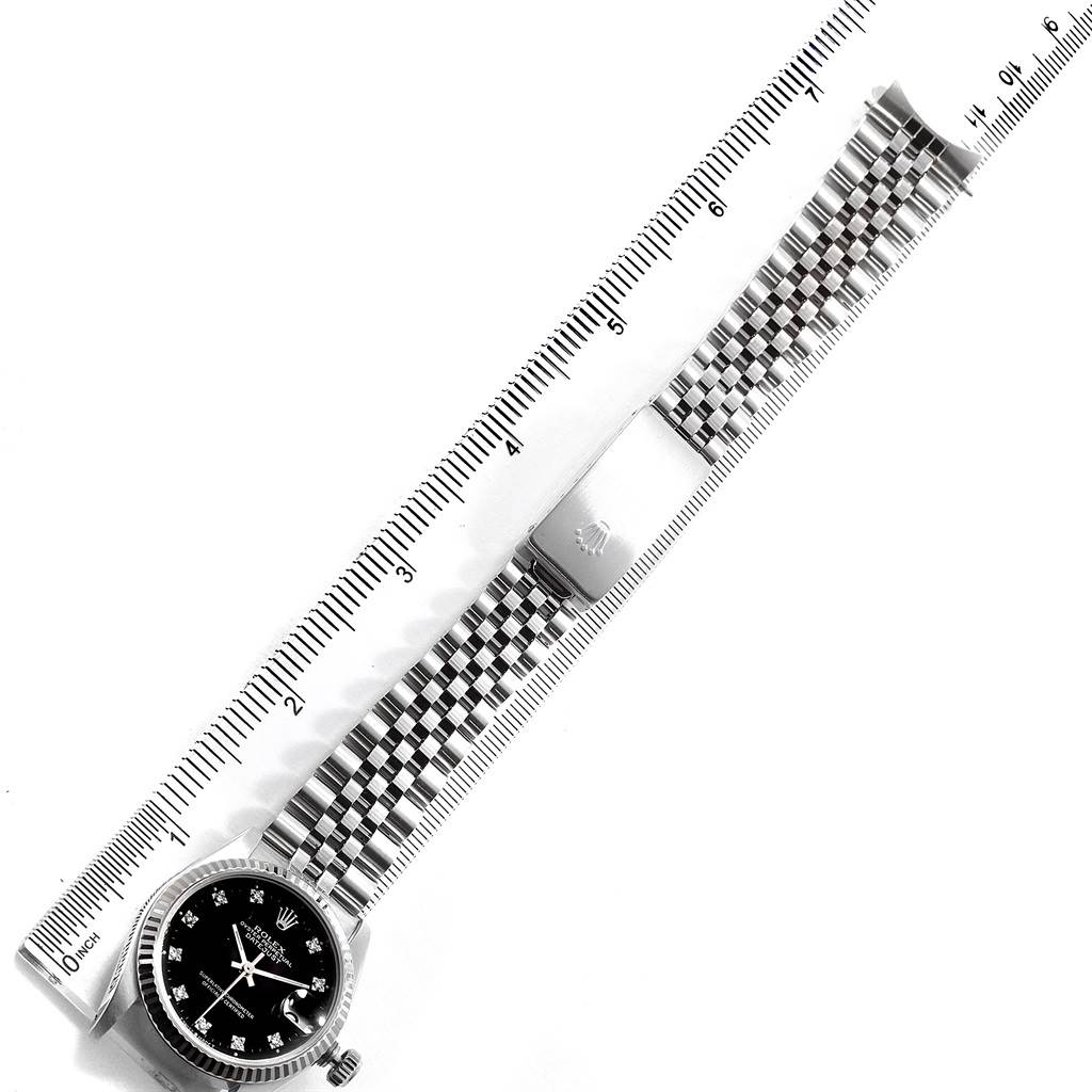 The image shows a top-down view of a mid-size Rolex watch with a black dial and jubilee bracelet beside a ruler for scale.