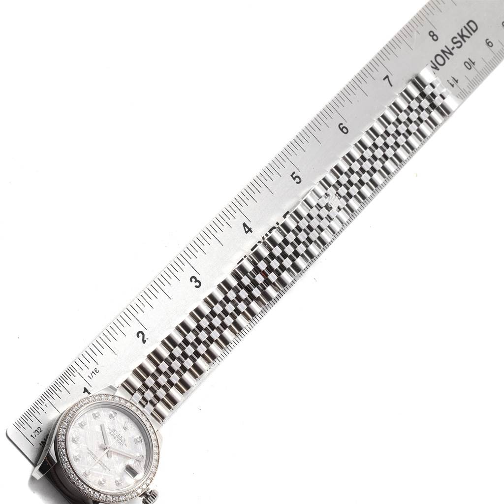 The image shows a Mid-Size Rolex watch lying flat alongside a metallic ruler, highlighting the bracelet and face from a top-down angle.