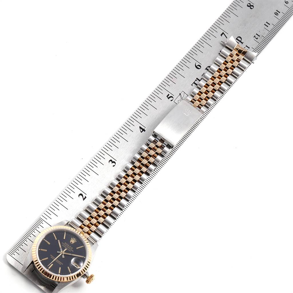 The image shows a Mid-Size Rolex watch laid flat with its black dial, gold bezel, and two-tone Jubilee bracelet extended and measured with a ruler.