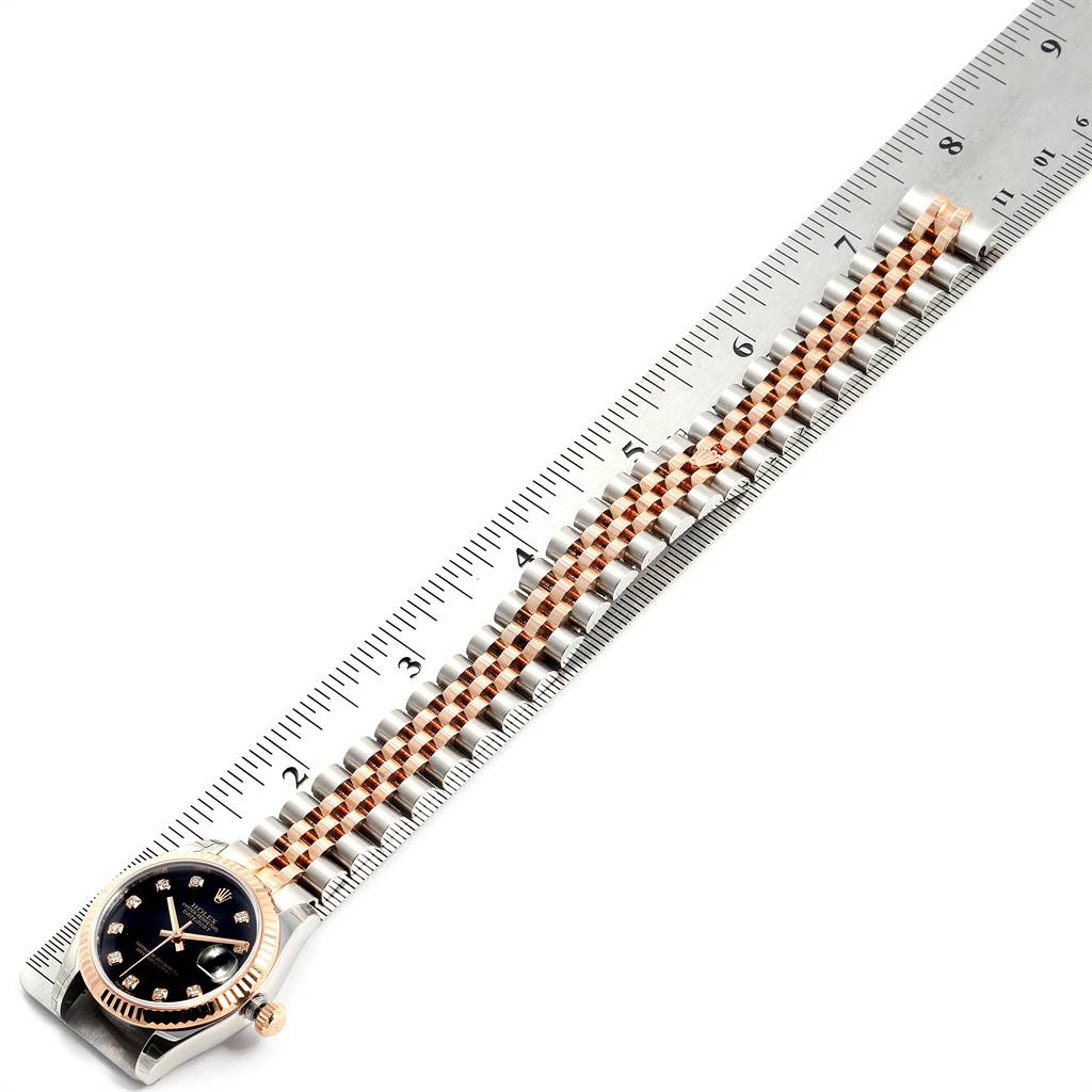 The image shows the top view of a Rolex Mid-Size 178271 Women's Steel and Gold (two tone) Black Dial 178271 Women's Steel and Gold (two tone) Black Dial watch with its black dial and bracelet laid out alongside a ruler.