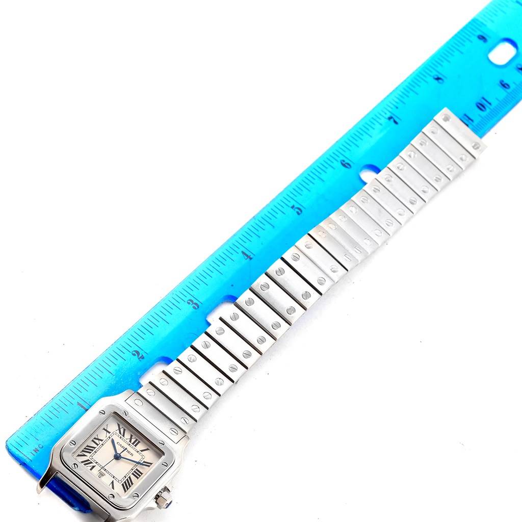 The image shows a top-down view of a Cartier Santos W20060D6 Men
s Stainless Steel Silver Dial W20060D6 Men
s Stainless Steel Silver Dial watch next to a blue ruler, displaying the watch
s face and band.