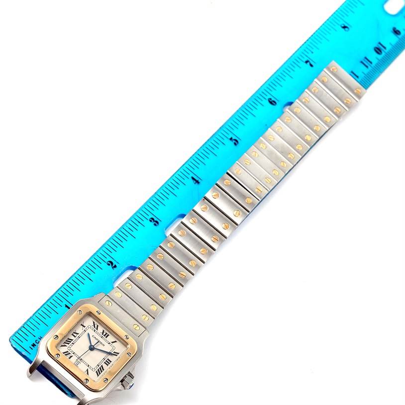 The Cartier Santos W20011C4 Stainless Steel Silver Dial W20011C4 Stainless Steel Silver Dial watch is shown from an angled top view with its watch face and entire band alongside a blue ruler.