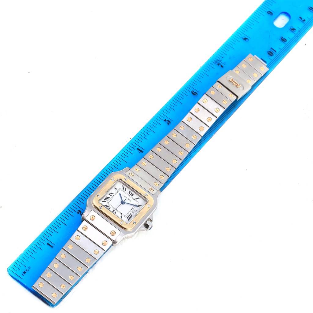 The image shows a top view of a Santos model Cartier watch, including its face and entire band, alongside a blue ruler.