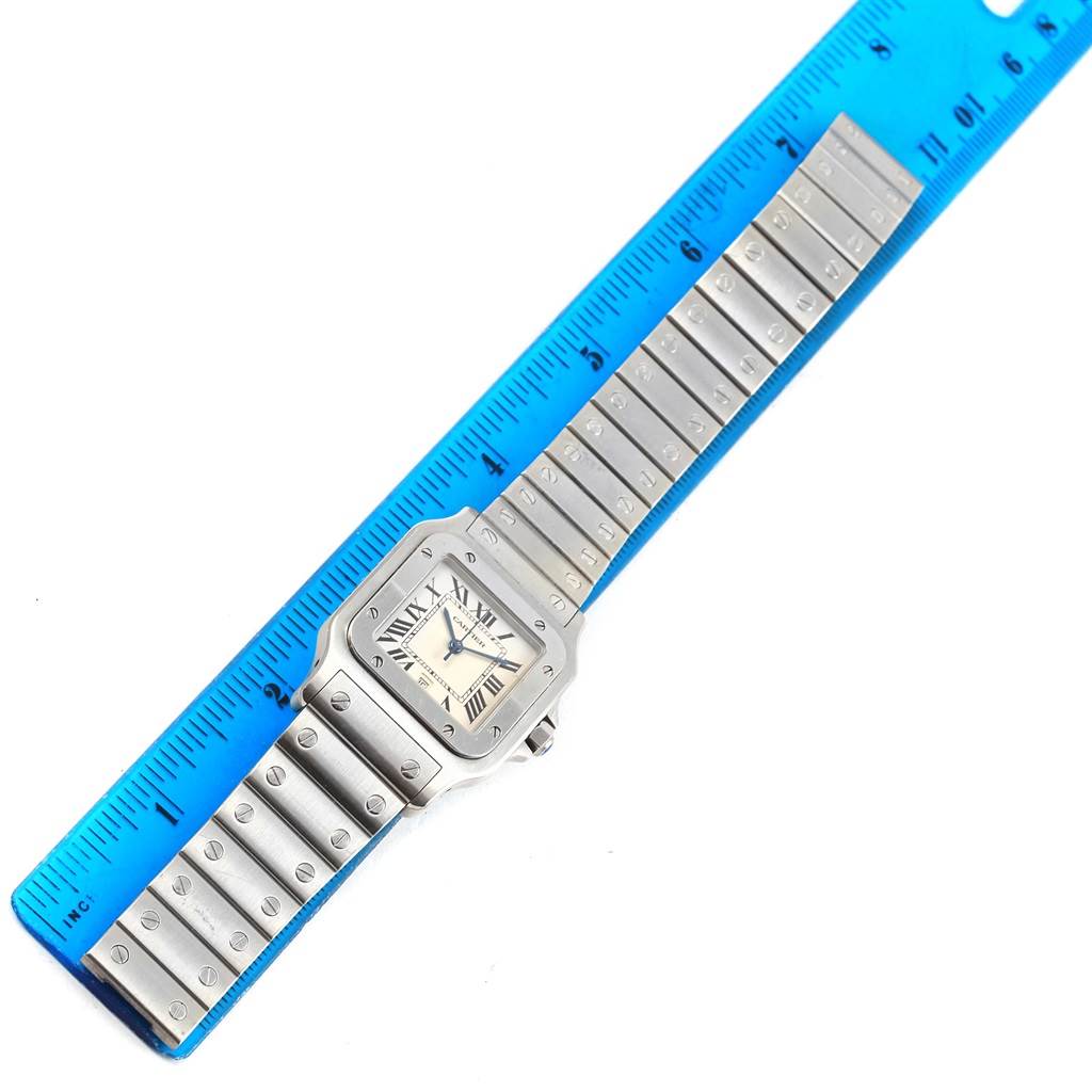 The image shows the front of the Cartier Santos W20060D6 Stainless Steel Silver Dial W20060D6 Stainless Steel Silver Dial watch with its bracelet, lying next to a blue ruler.