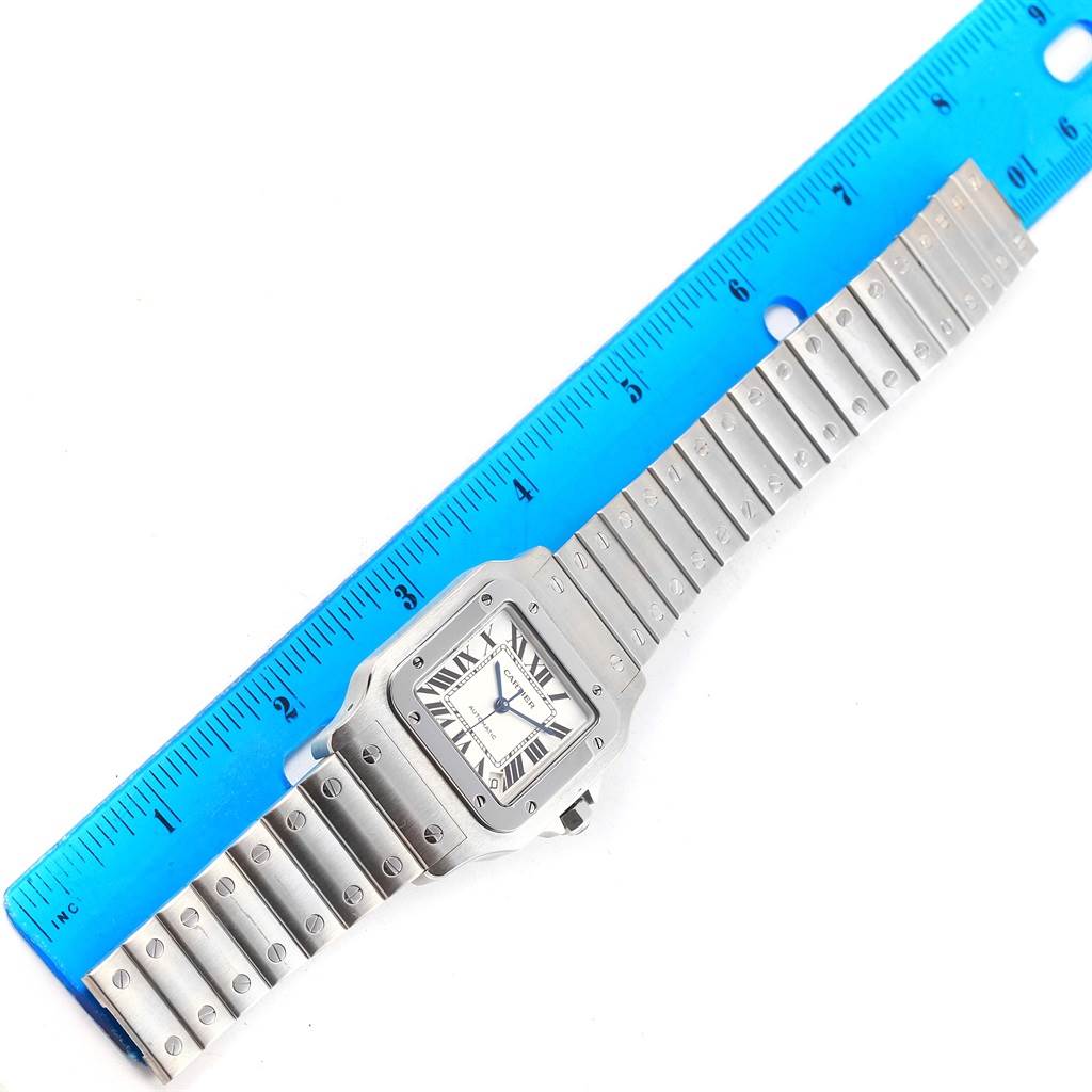 The Cartier Santos W20098D6 Men's Stainless Steel Silver Dial W20098D6 Men's Stainless Steel Silver Dial watch is shown from a top-down angle, displaying its face, bezel, and metal bracelet alongside a blue ruler.
