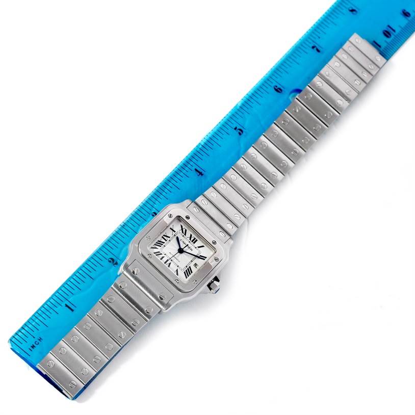 The Cartier Santos W20055D6 Men
s Stainless Steel Silver Dial W20055D6 Men
s Stainless Steel Silver Dial watch is shown from a top-down angle, displaying the full bracelet and face of the watch alongside a blue ruler.