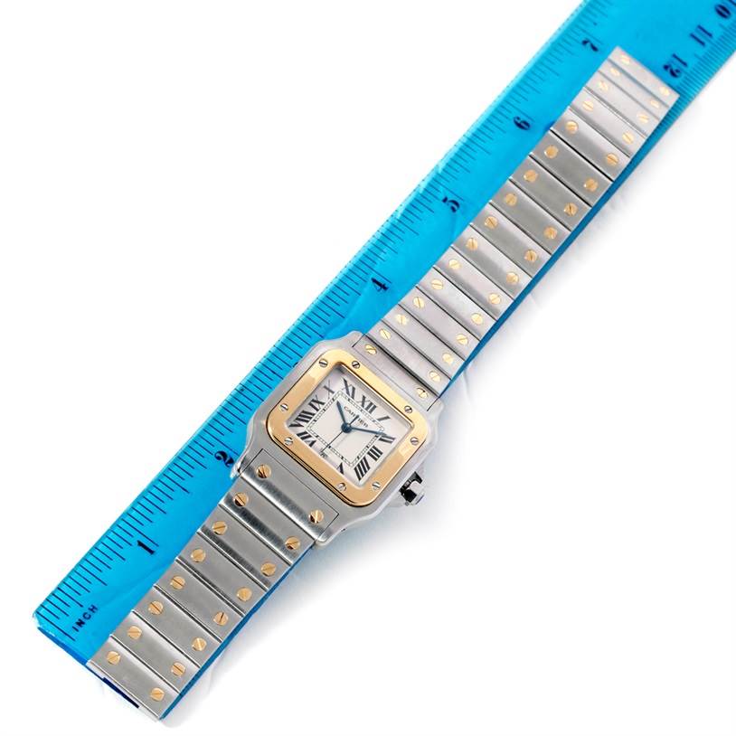 The image shows a Santos model by Cartier, featuring the watch face, bezel, and bracelet, alongside a blue ruler.