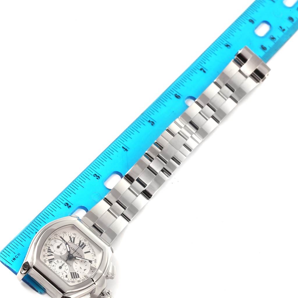 The image shows a close-up top view of a Cartier Roadster W62006X6 Men's Stainless Steel Silver Dial W62006X6 Men's Stainless Steel Silver Dial watch with a metal bracelet alongside a blue ruler for size reference.