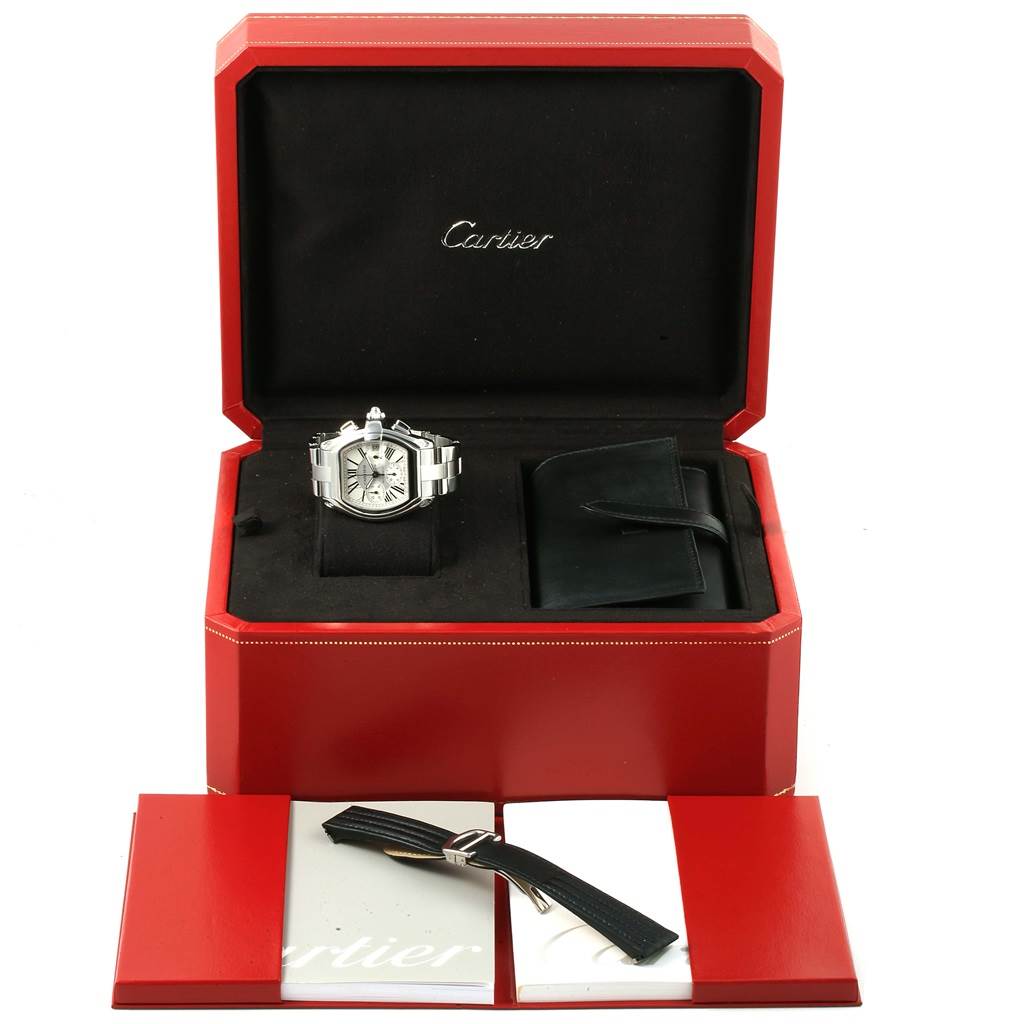 The image shows a Cartier Roadster W62019X6 Men's Stainless Steel Silver Dial W62019X6 Men's Stainless Steel Silver Dial watch in a red box with a spare strap, a booklet, and a pouch.