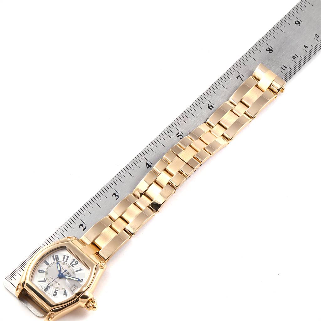 The image shows a Cartier Roadster W62005V1 Men's Yellow Gold Silver Dial W62005V1 Men's Yellow Gold Silver Dial watch placed on a ruler, viewed from the top, showcasing its face and band.