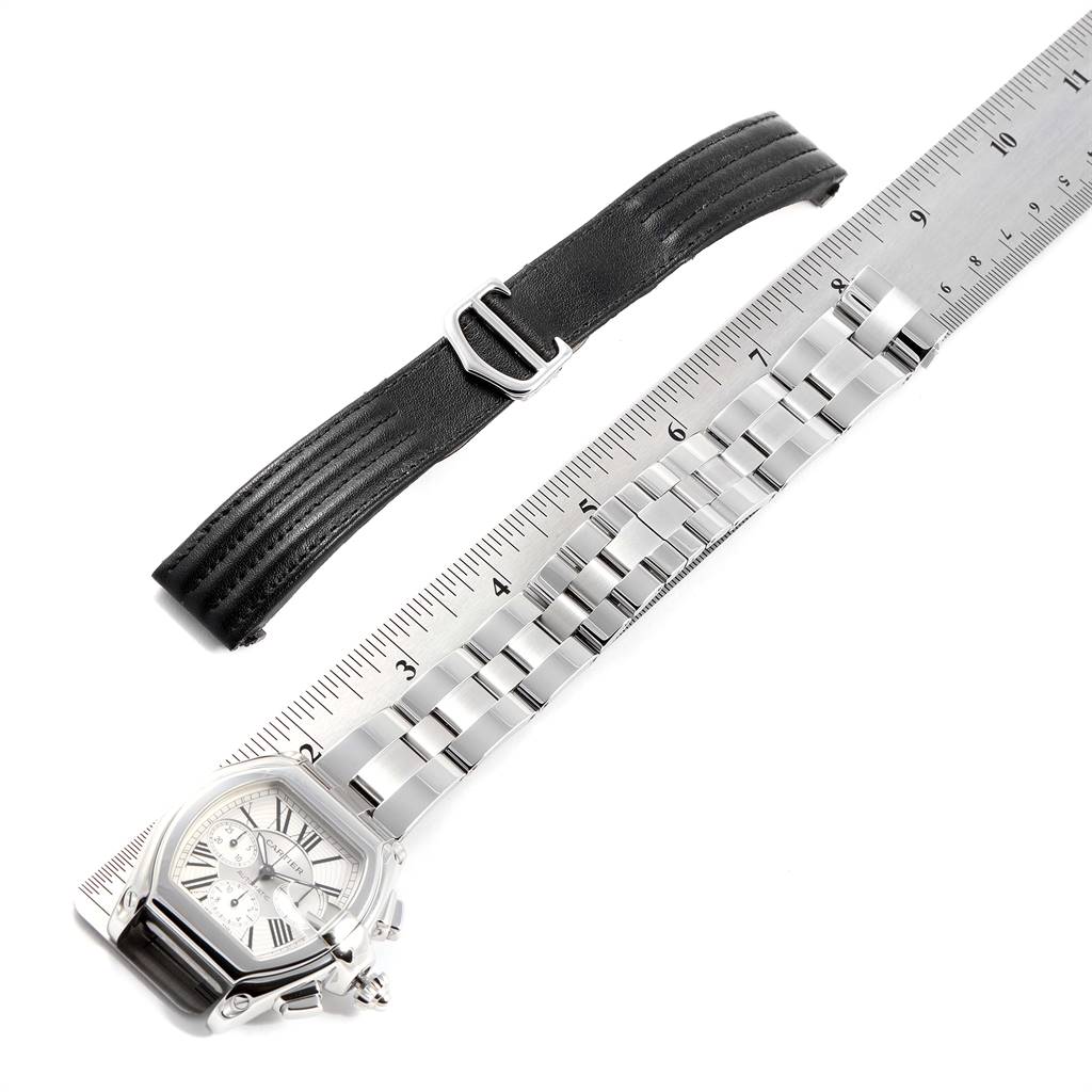 The image shows a top-down view of a Cartier Roadster W62019X6 Men
s Stainless Steel Silver Dial W62019X6 Men
s Stainless Steel Silver Dial watch with a metal bracelet and a black leather strap alongside a ruler.