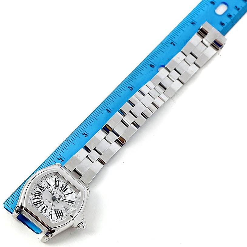 The image shows a top-down view of a Cartier Roadster W62032X6 Men
s Stainless Steel Silver Dial W62032X6 Men
s Stainless Steel Silver Dial watch with its face, bezel, and metal bracelet alongside a blue ruler.