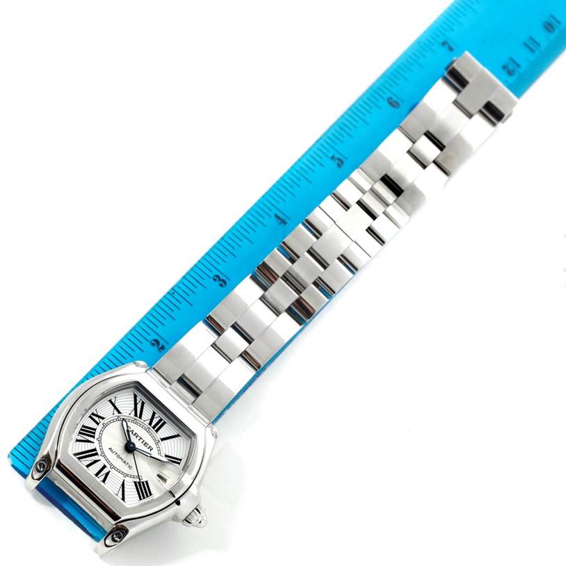 The image shows the Cartier Roadster W62025V3 Men
s Stainless Steel Silver Dial W62025V3 Men
s Stainless Steel Silver Dial watch diagonally positioned next to a blue ruler, highlighting its face and bracelet.