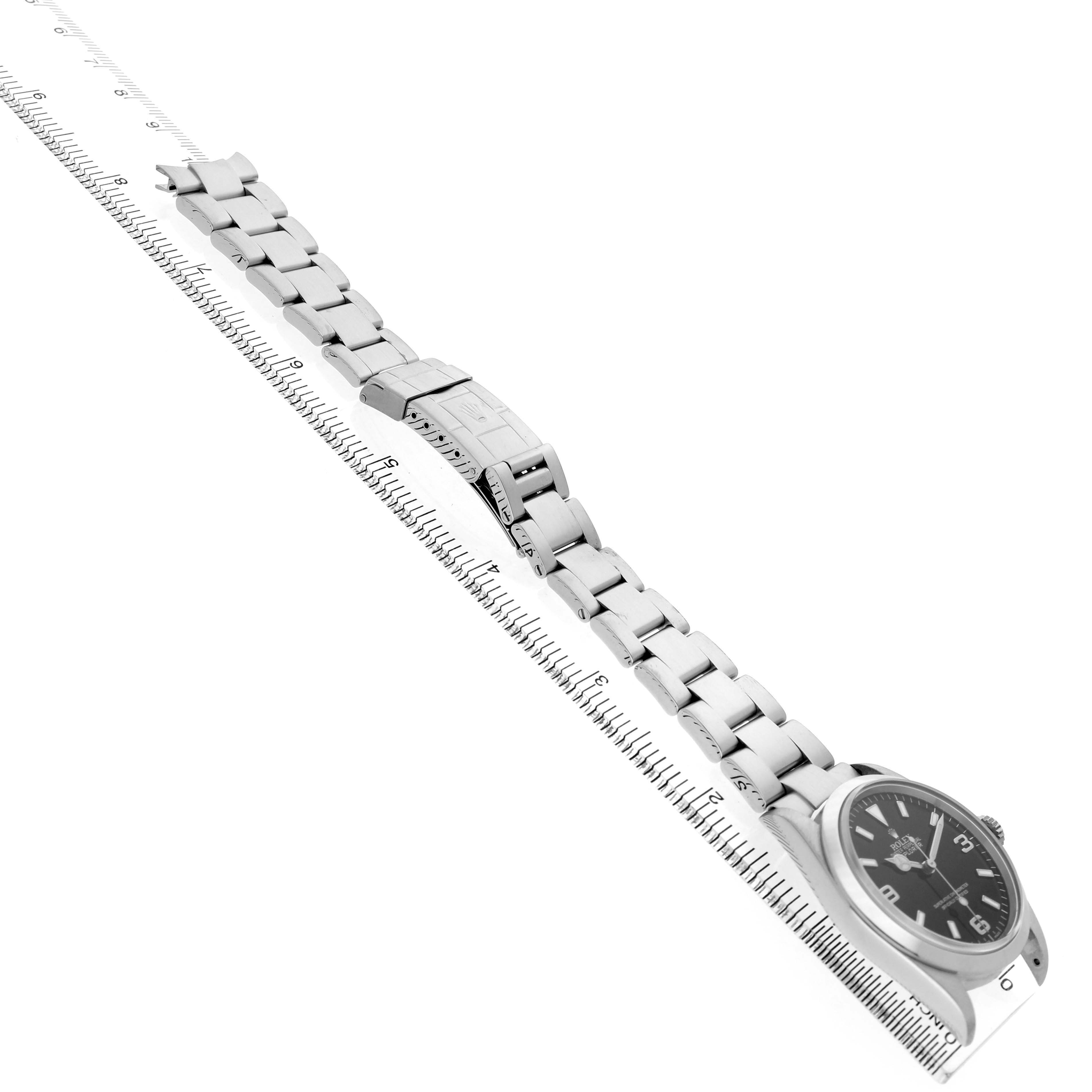 The Rolex Explorer 114270 Men's Stainless Steel Black Dial watch is shown at an angle displaying the bracelet and clasp, alongside a measuring ruler.
