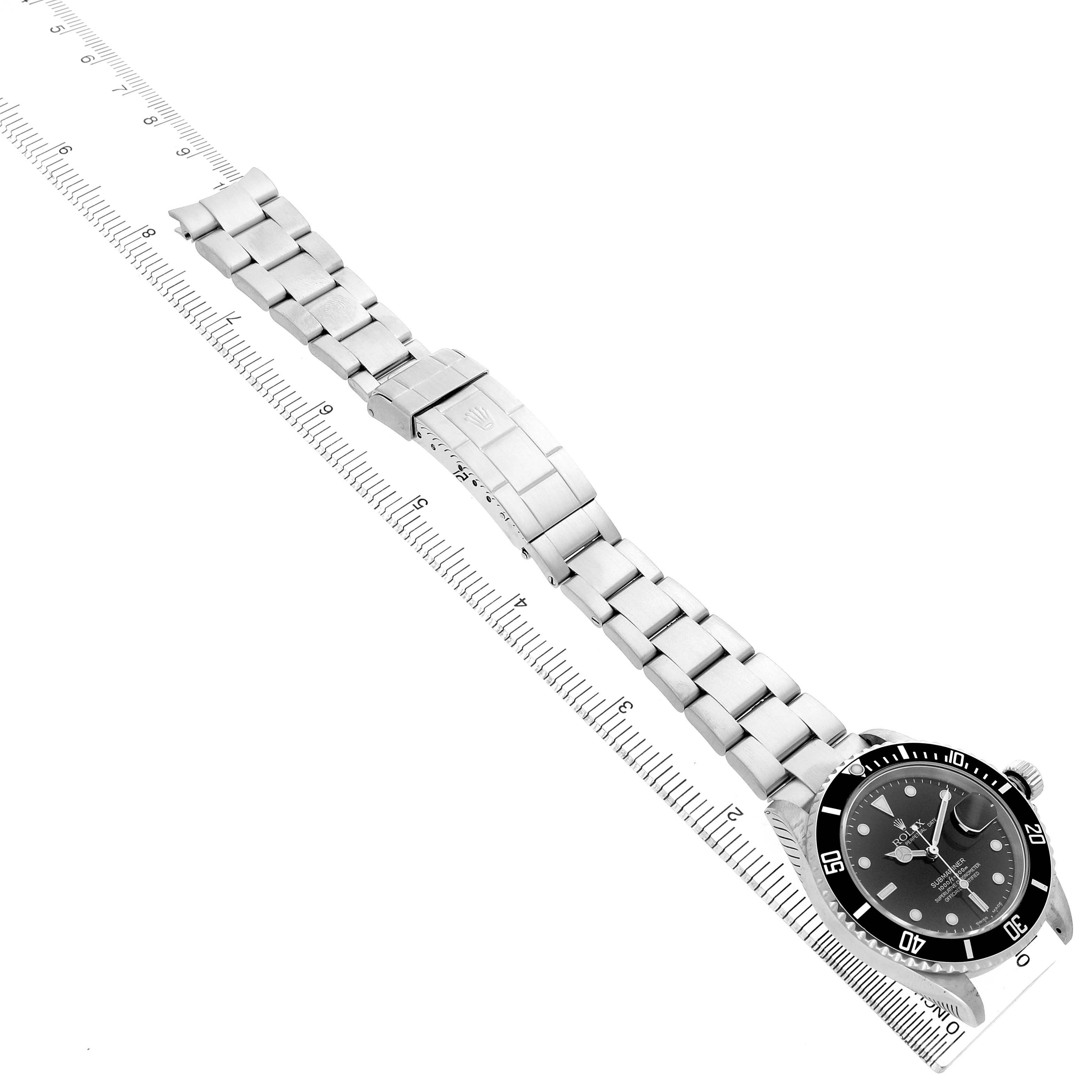 The Rolex Submariner 16610 Men's Stainless Steel Black Dial watch is shown from an overhead angle, displaying its face, bezel, and metal bracelet alongside a ruler.