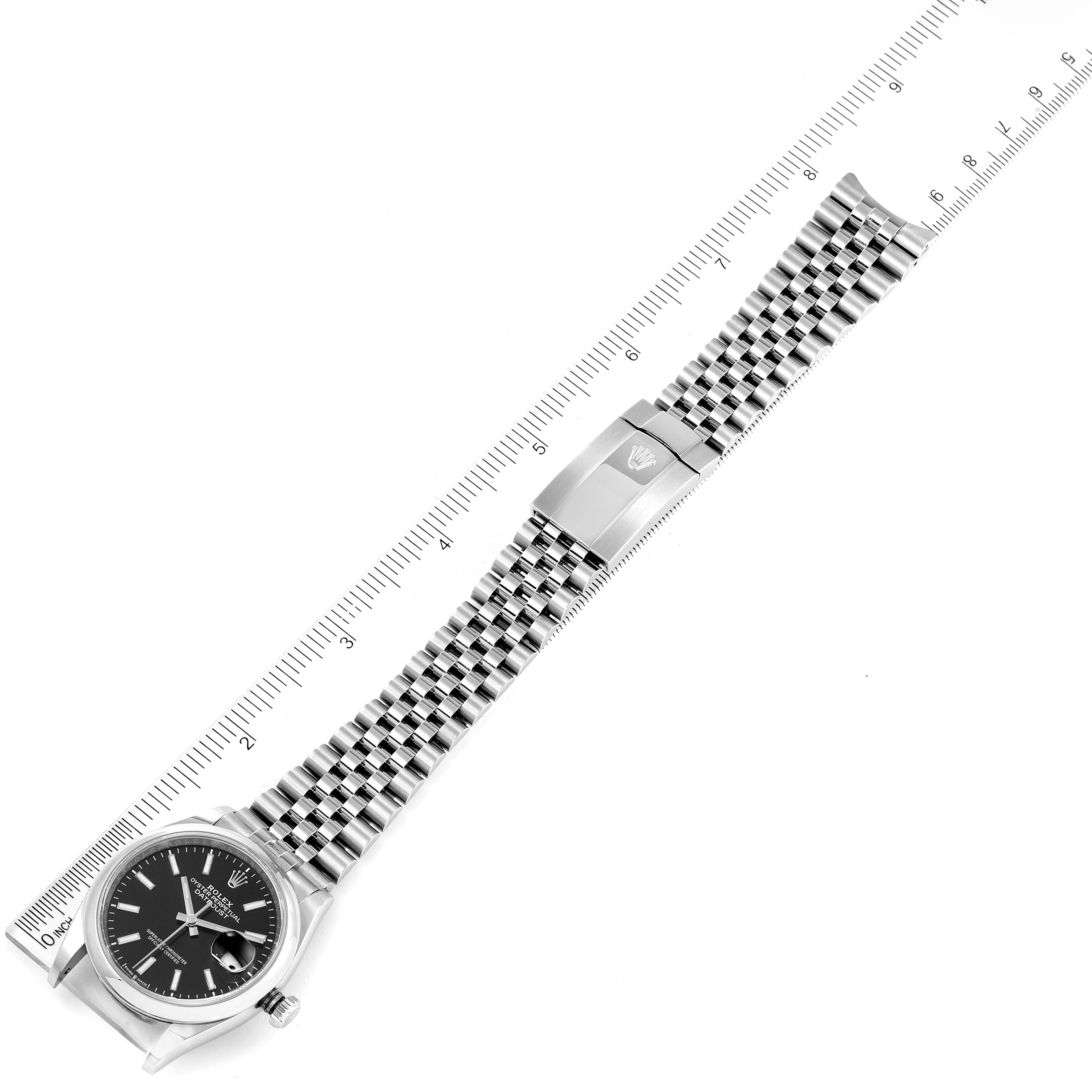 The image shows a top-down view of a Rolex Datejust 126200 Men
s Stainless Steel Black Dial 126200 Men
s Stainless Steel Black Dial watch, highlighting its face, bracelet, and clasp alongside a ruler for scale.