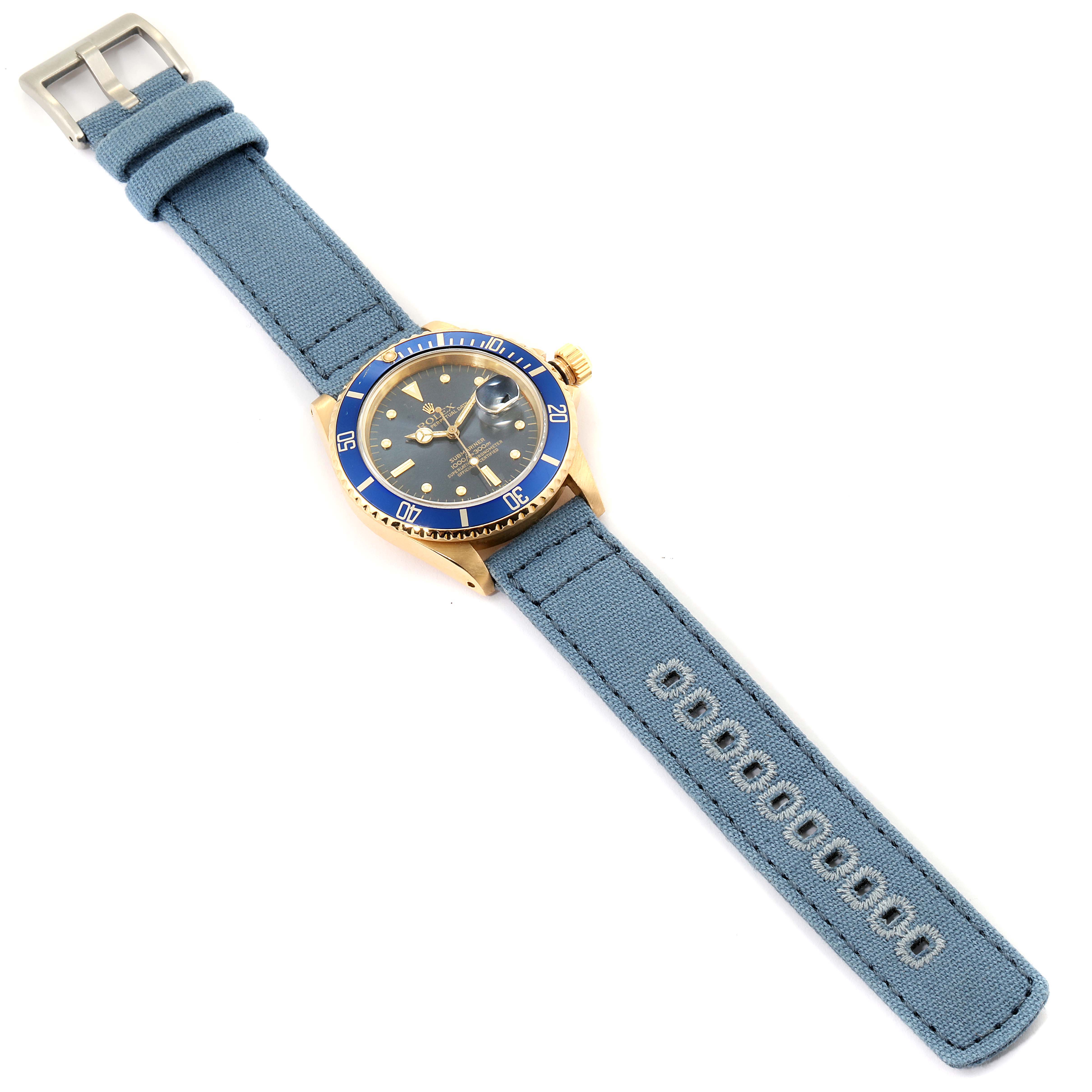 The image shows a Rolex Submariner 16808 Men
s Yellow Gold Blue Dial 16808 Men
s Yellow Gold Blue Dial watch with a blue bezel, blue dial, and grey fabric strap at an aerial view.