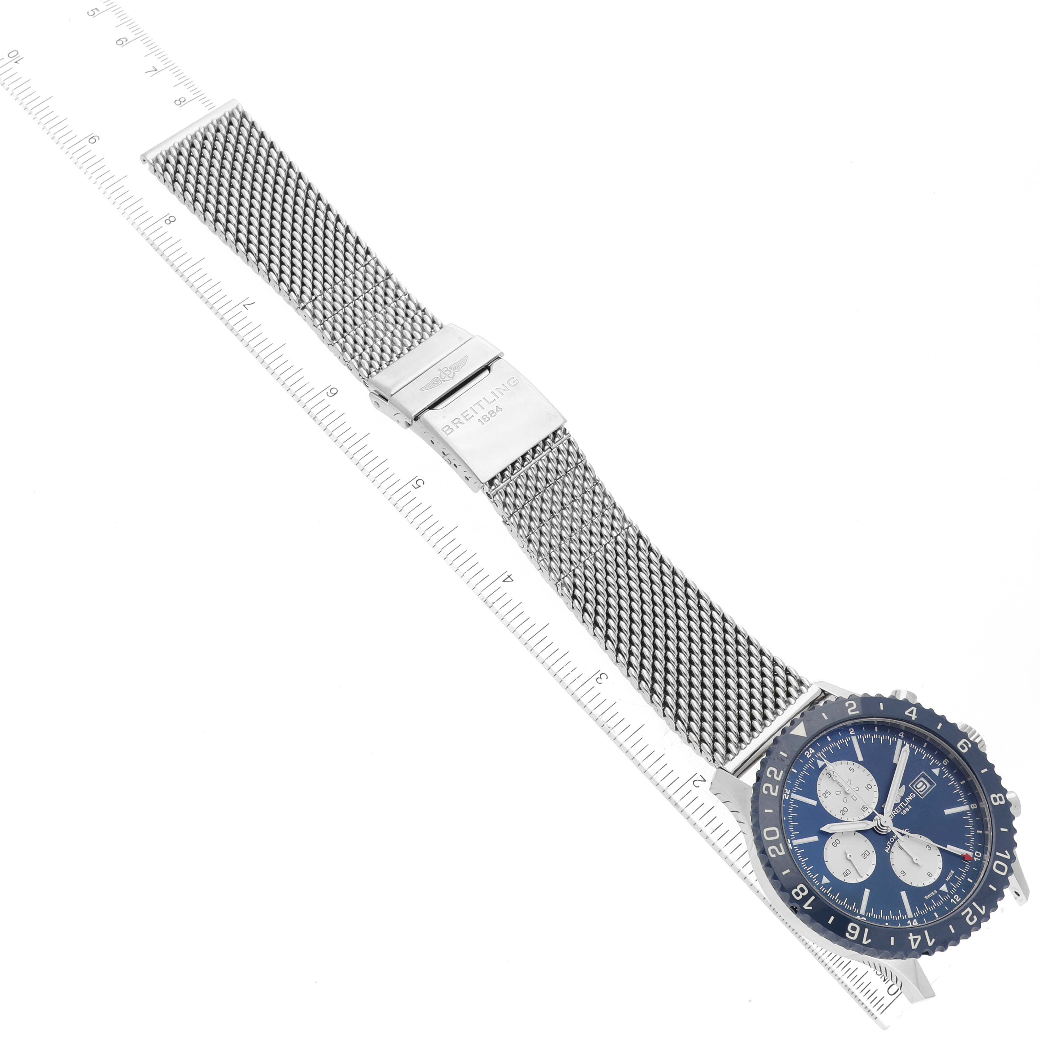 The image shows a top view of a Breitling Chronoliner Y243106/C970 Men's Stainless Steel Blue Dial Y243106/C970 Men's Stainless Steel Blue Dial watch, highlighting its face, bezel, and metal mesh strap against a ruler scale.