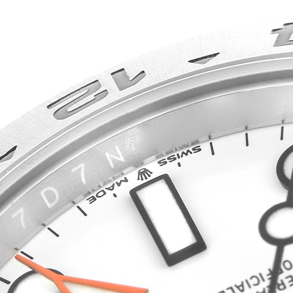 The image shows a close-up of the Rolex Explorer 226570 Men
s Stainless Steel White Dial 226570 Men
s Stainless Steel White Dial
s bezel, part of the dial, and part of an orange hand.