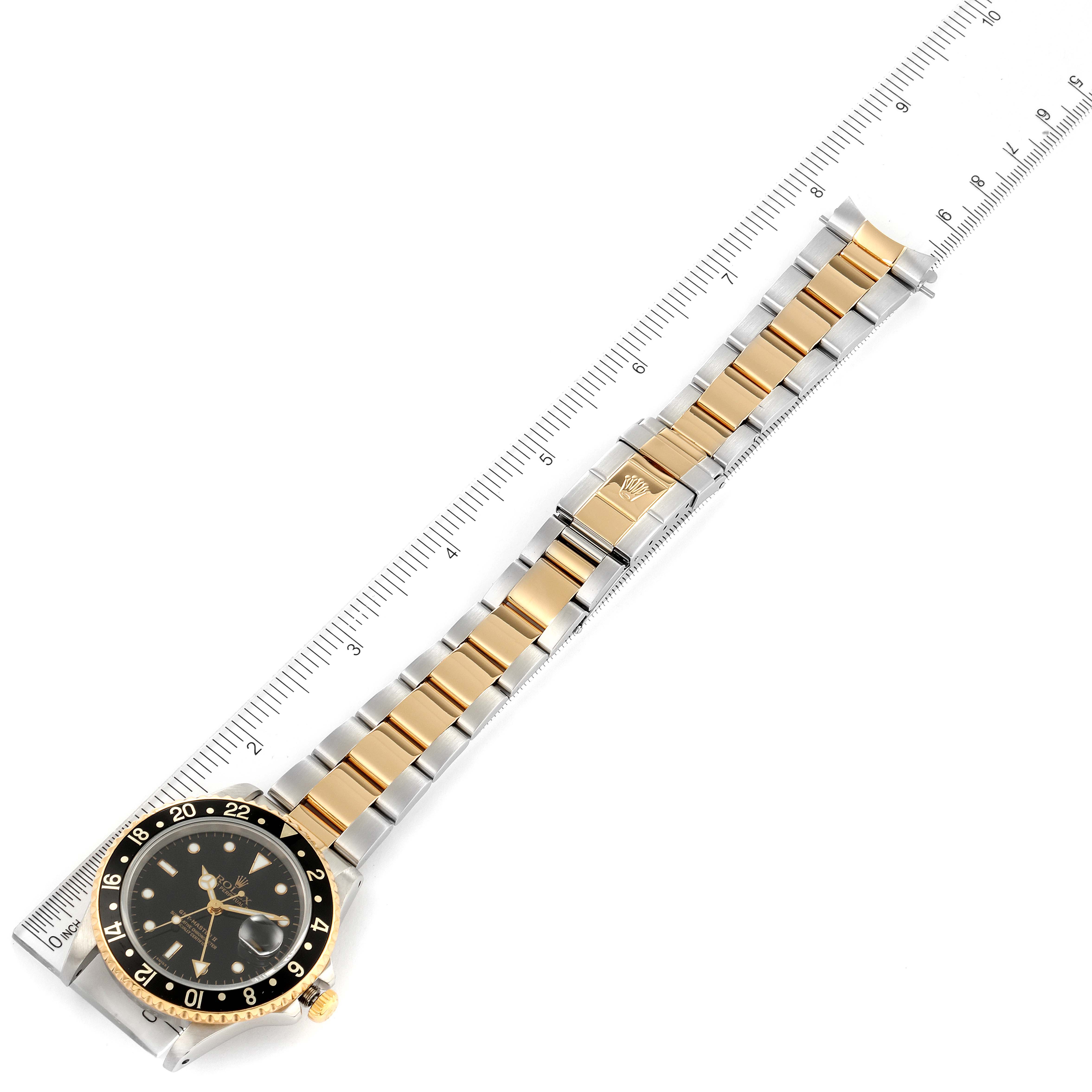 The image shows a top view of a Rolex GMT-Master 16713 Men's Steel and Gold (two tone) Black Dial 16713 Men's Steel and Gold (two tone) Black Dial watch, displaying the face, bezel, and two-tone bracelet next to a ruler.
