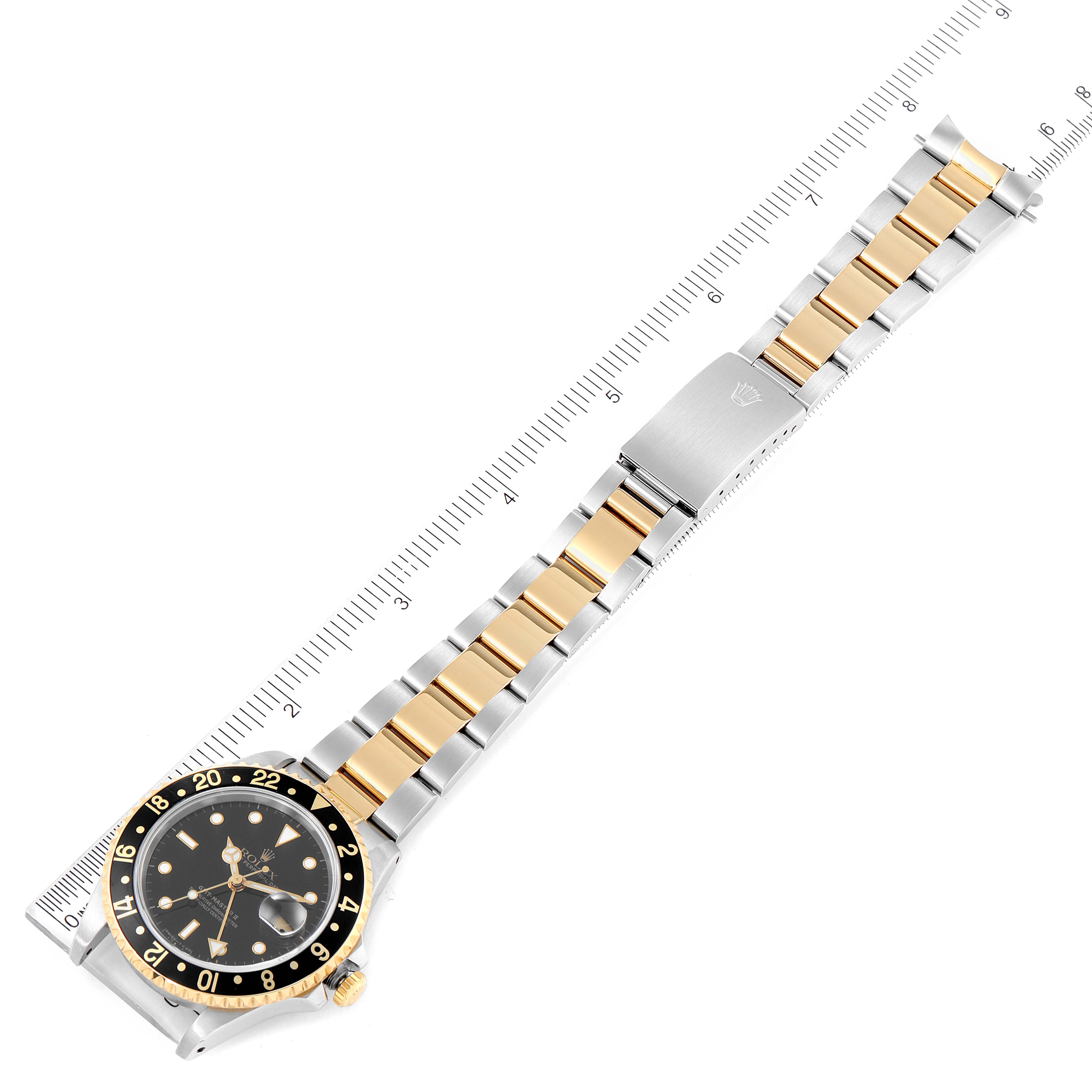 The Rolex GMT-Master 16713 Men's Steel and Gold (two tone) Black Dial 16713 Men's Steel and Gold (two tone) Black Dial watch is displayed flat, showing its face, bezel, and metal bracelet with a ruler for a size reference.