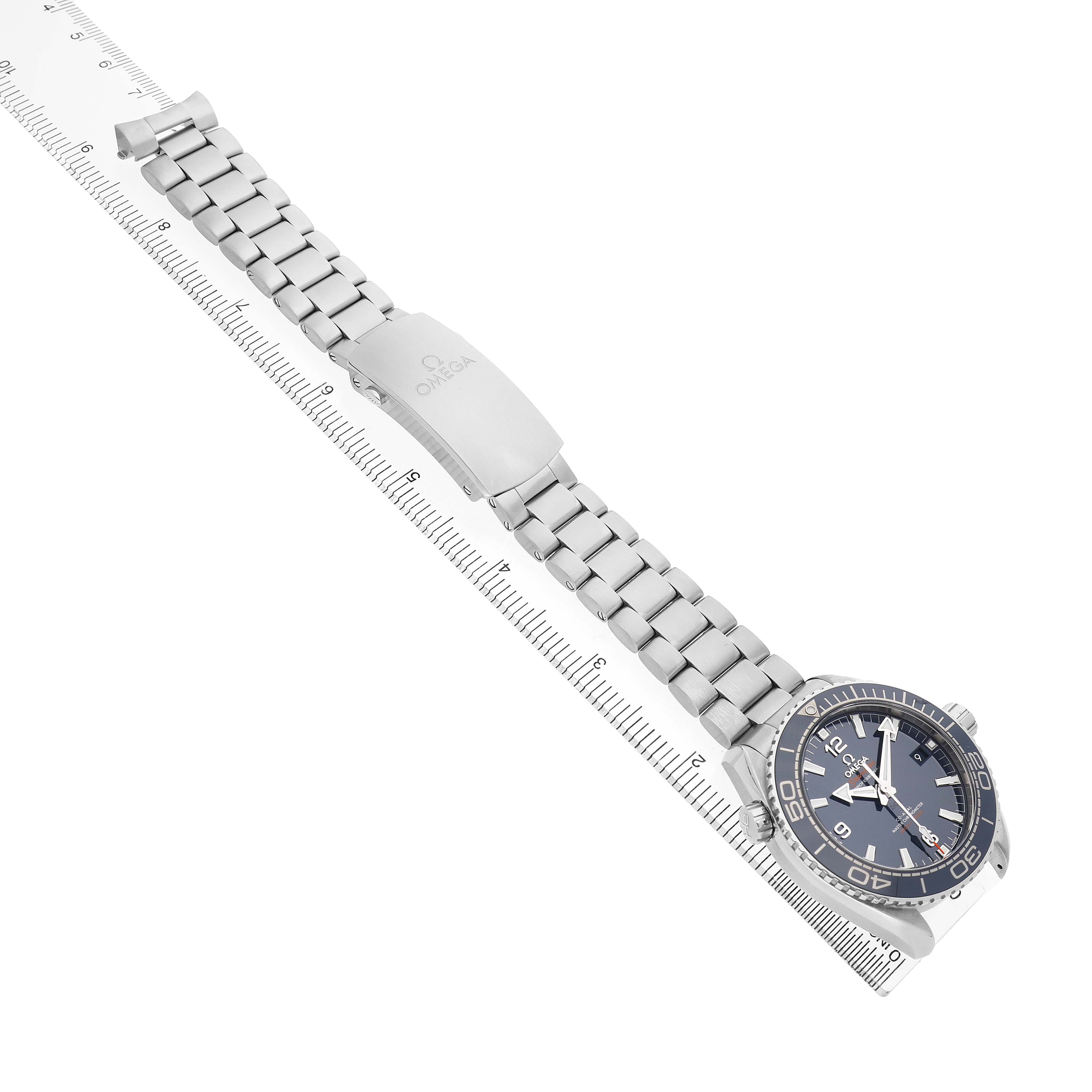 The Omega Planet Ocean 215.30.44.21.03.001 Men's Stainless Steel Blue Dial watch is shown at an angle displaying the metal bracelet, clasp, and face, positioned diagonally on a ruler.
