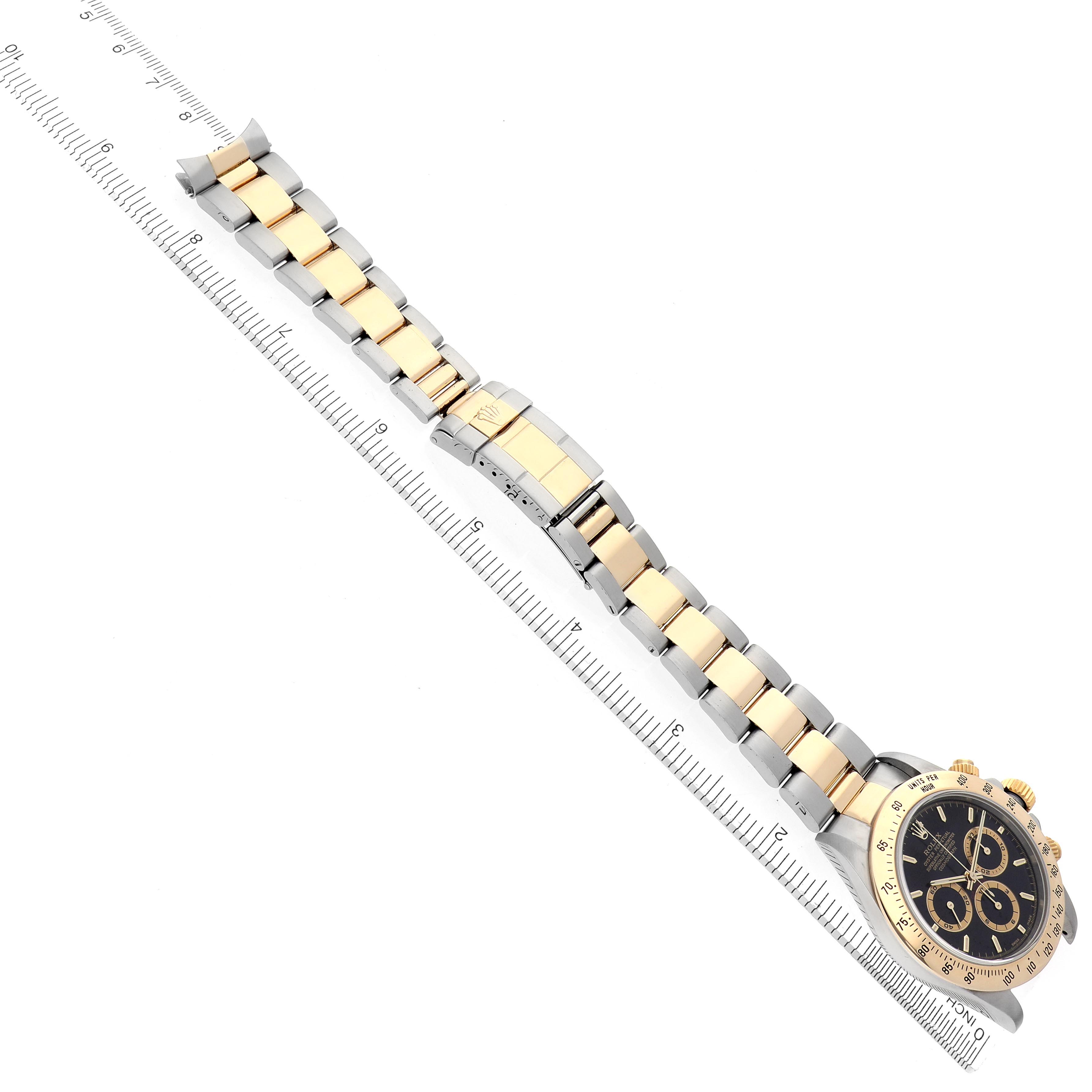 The image shows a Rolex Daytona 16523 Men's Steel and Gold (two tone) Black Dial 16523 Men's Steel and Gold (two tone) Black Dial watch from a top-down angle, highlighting the full bracelet and watch face alongside a ruler for scale.