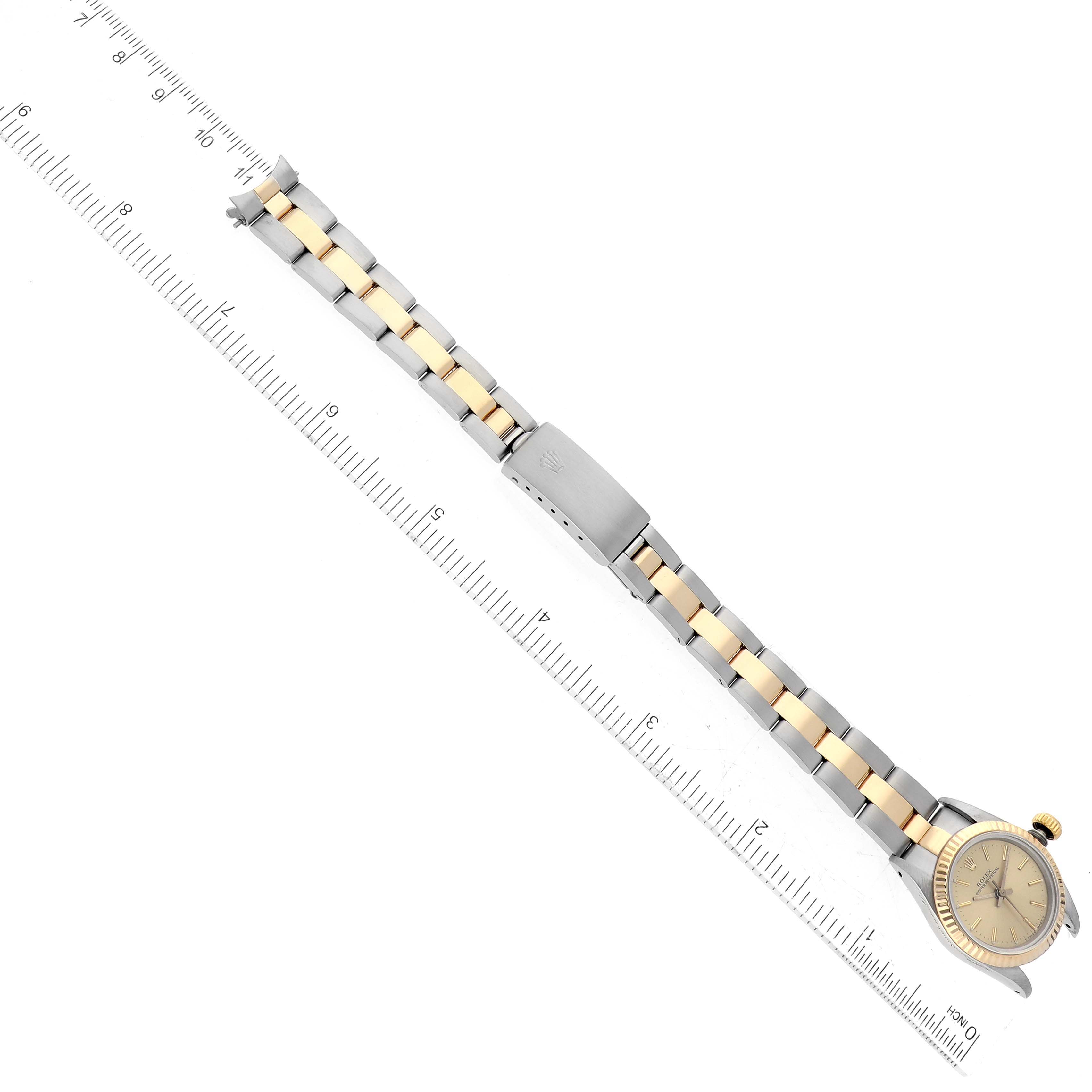 This image shows a top-down view of a Rolex Oyster Perpetual 67193 Women's Steel and Gold (two tone) Champagne Dial 67193 Women's Steel and Gold (two tone) Champagne Dial watch displaying its face, crown, bracelet, and clasp next to a ruler.