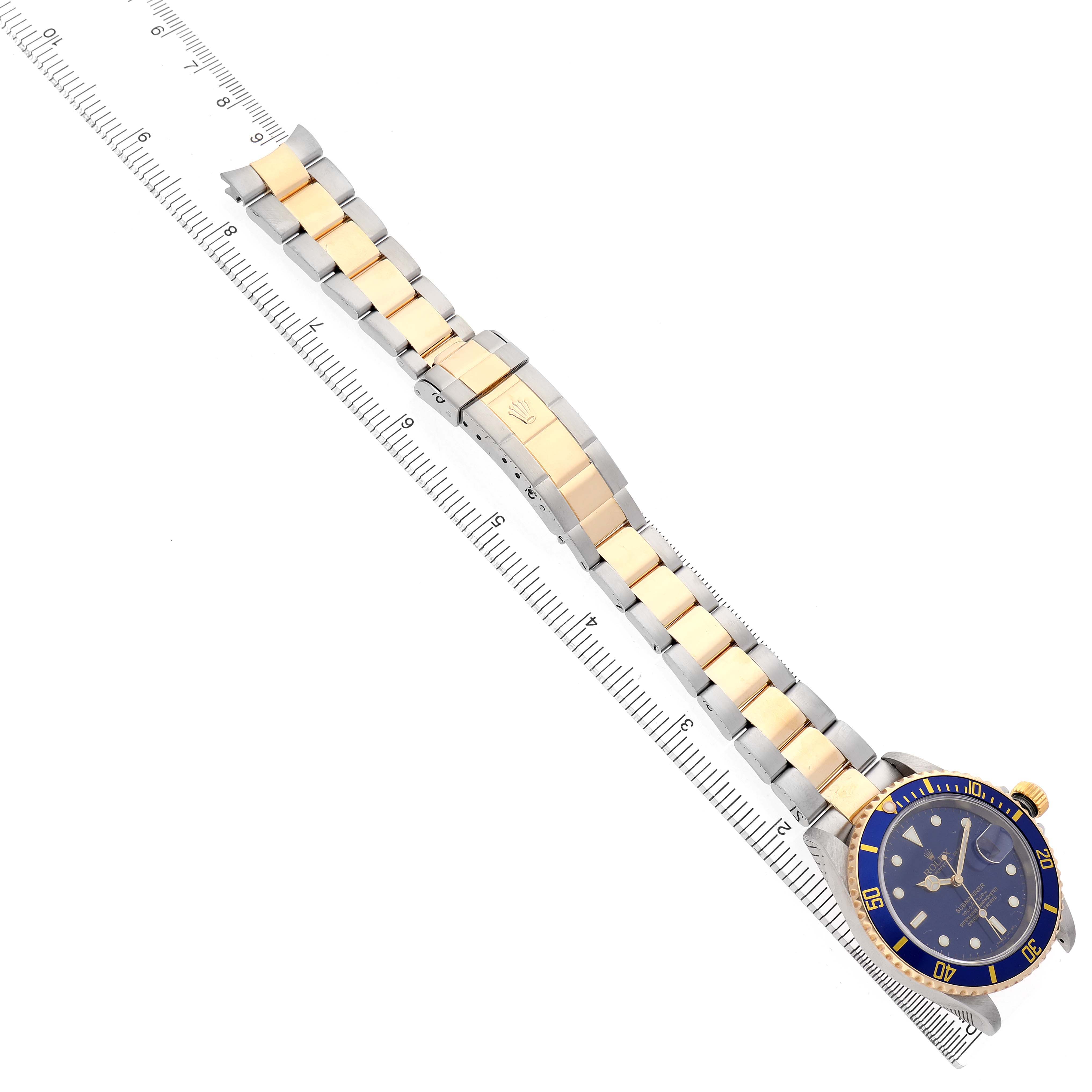 The Rolex Submariner 16613 Men's Steel and Gold (two tone) Blue Dial watch is shown diagonally, displaying its face, bezel, and full bracelet alongside a ruler for scale.
