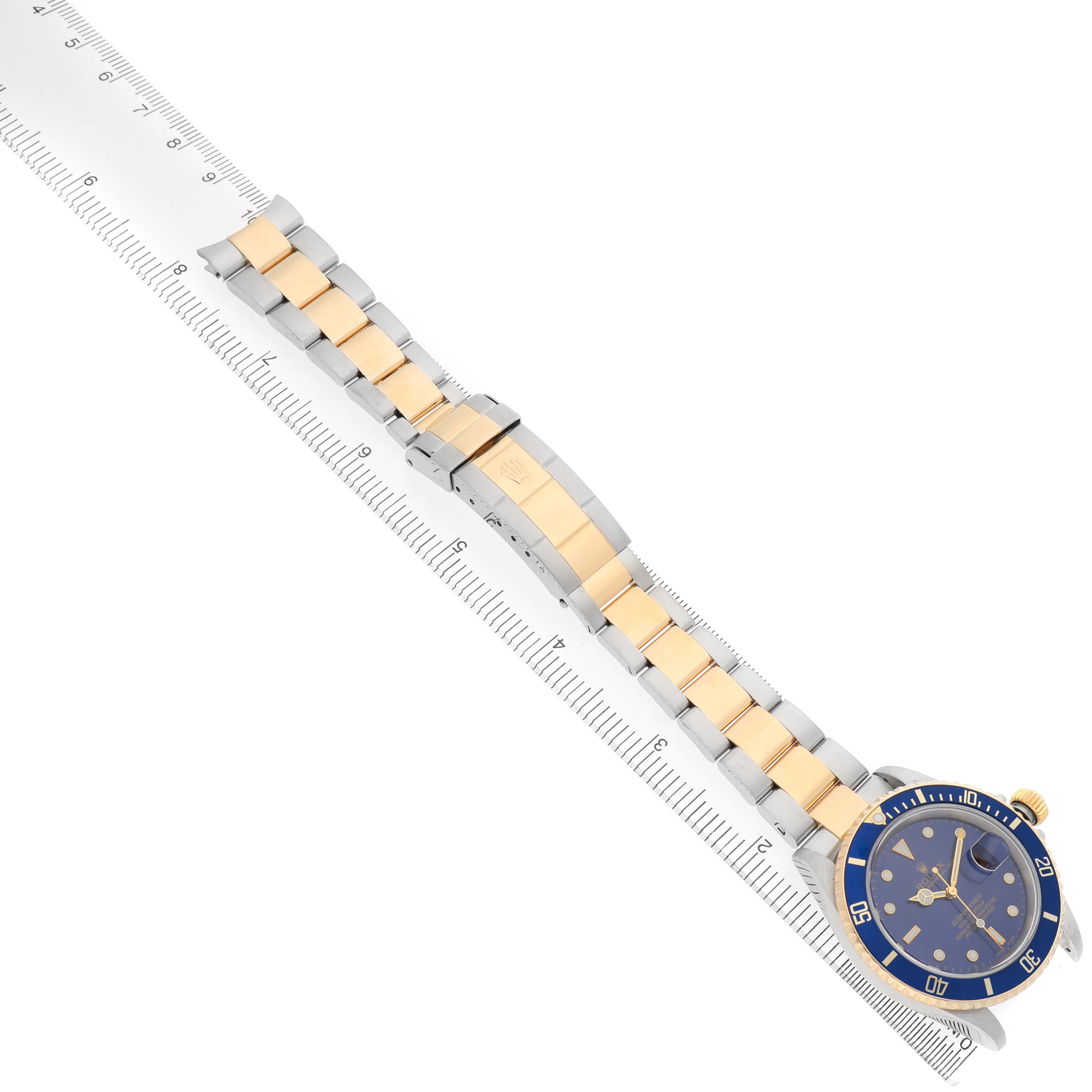 The Rolex Submariner 16613 Men's Steel and Gold (two tone) Blue Dial watch is shown from a top-down angle, displaying the blue dial and two-tone bracelet alongside a ruler.