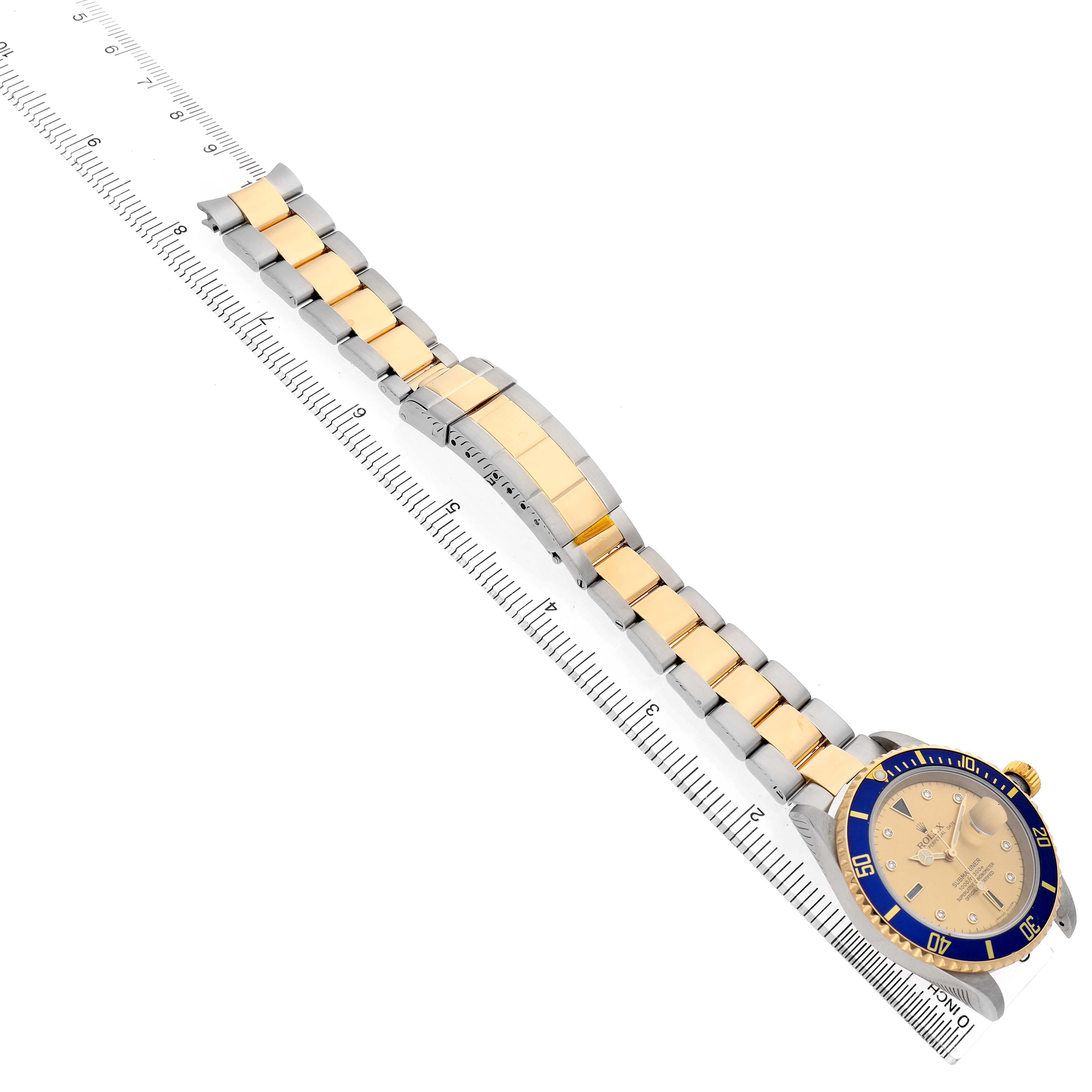 The Rolex Submariner 16613 Men's Steel and Gold (two tone) Champagne Dial watch is shown at an angled view, displaying the bracelet, clasp, and dial, placed alongside a ruler.