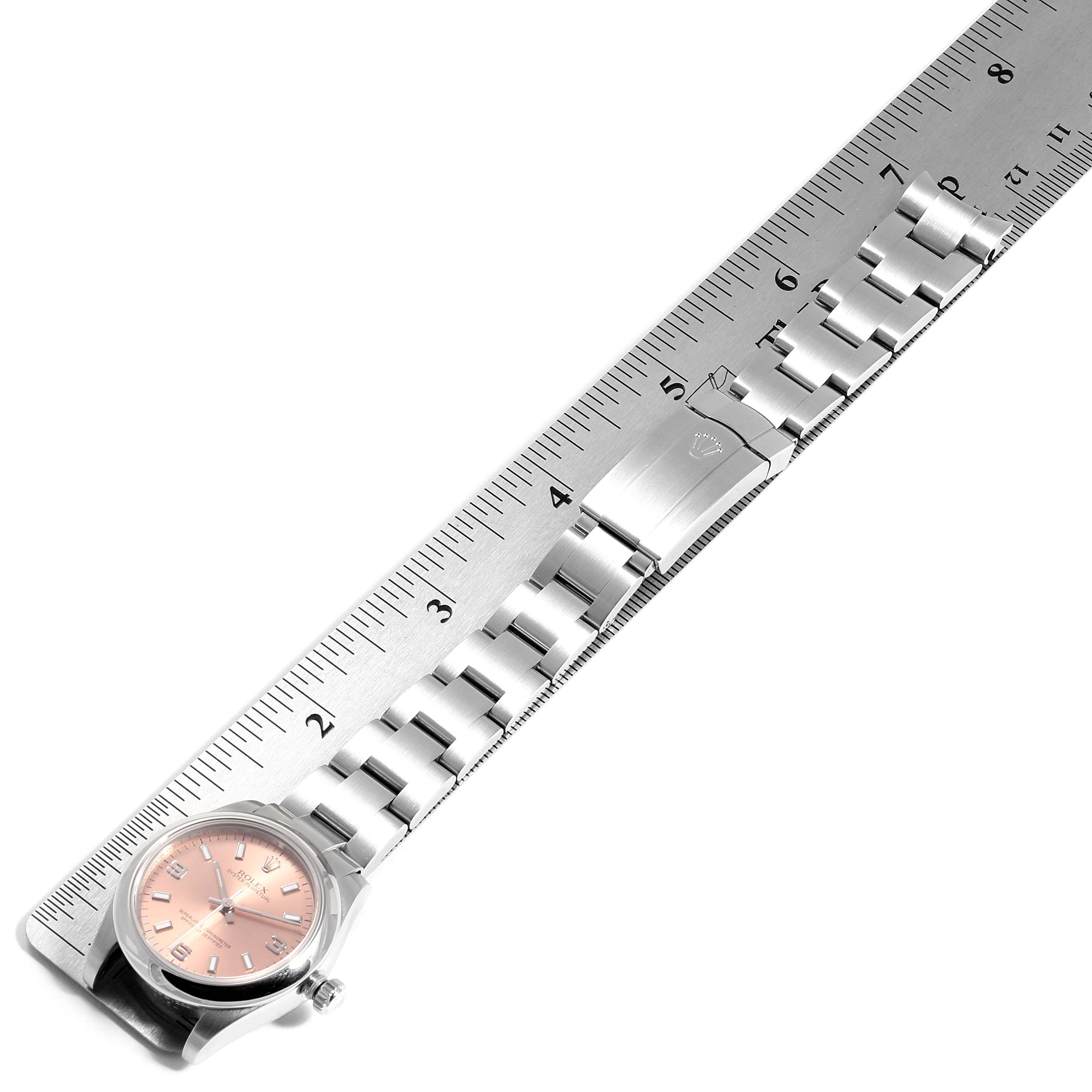 The image shows the front of a Rolex Mid-Size 177200 Women
s Stainless Steel Pink Dial 177200 Women
s Stainless Steel Pink Dial model watch with a pink dial and part of its stainless steel bracelet laid next to a ruler.