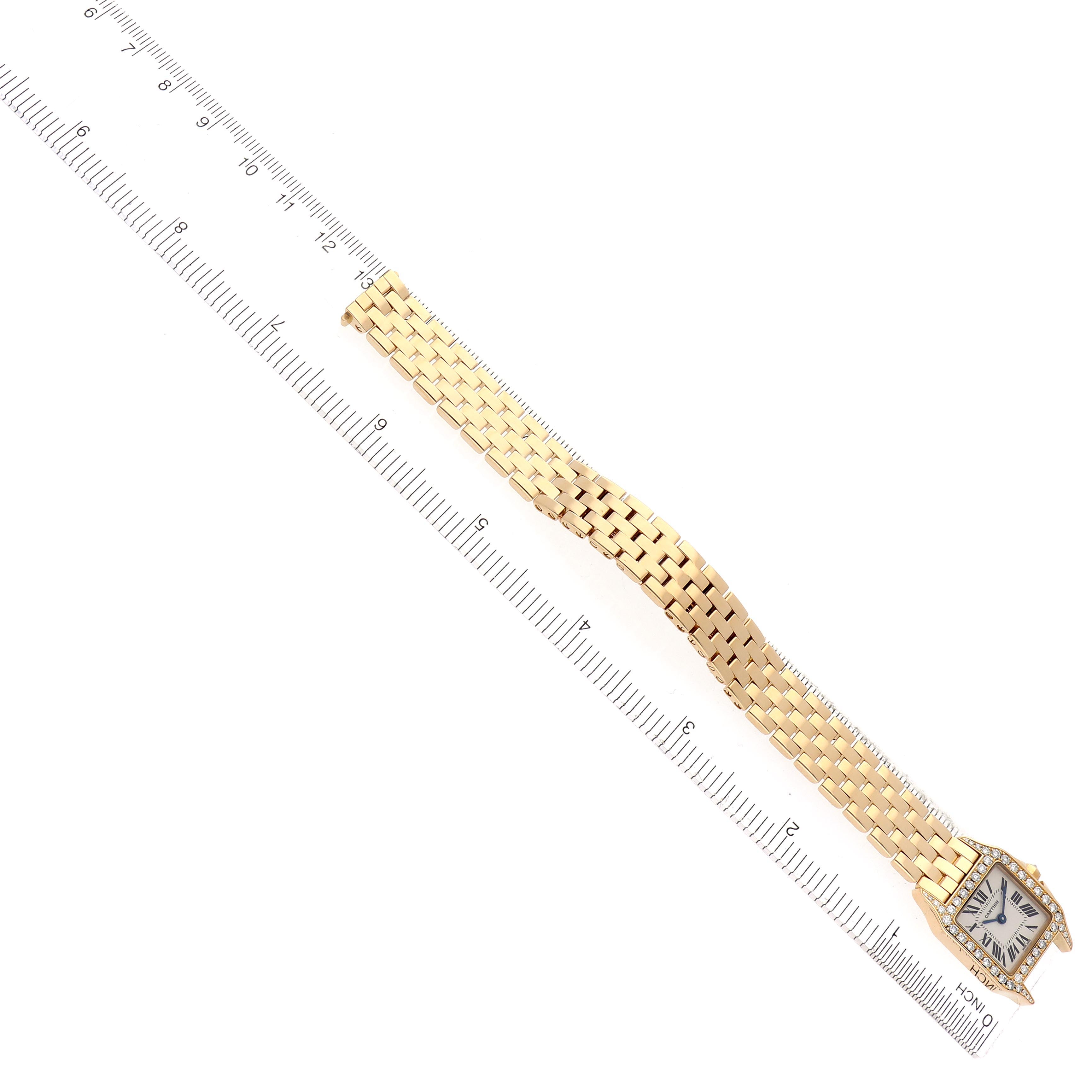 The image shows a Cartier Santos Demoiselle WF9001Y7 Women
s Yellow Gold Silver Dial WF9001Y7 Women
s Yellow Gold Silver Dial watch laid flat, displaying the gold bracelet and square face next to a ruler.