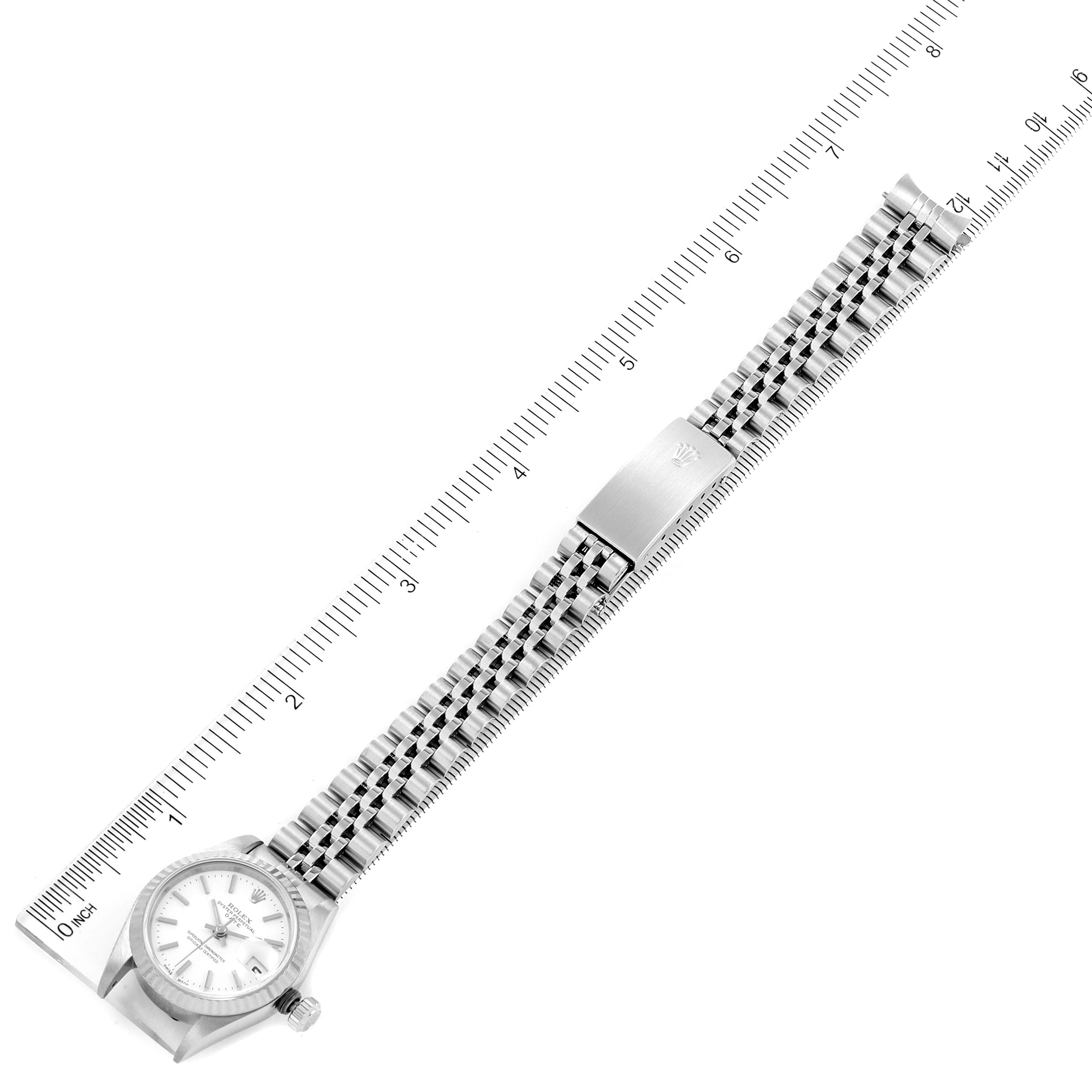 The image shows a Rolex Date 69174 Women
s Stainless Steel White Dial 69174 Women
s Stainless Steel White Dial watch from a top view, displaying the face, crown, bracelet, and clasp next to a ruler.