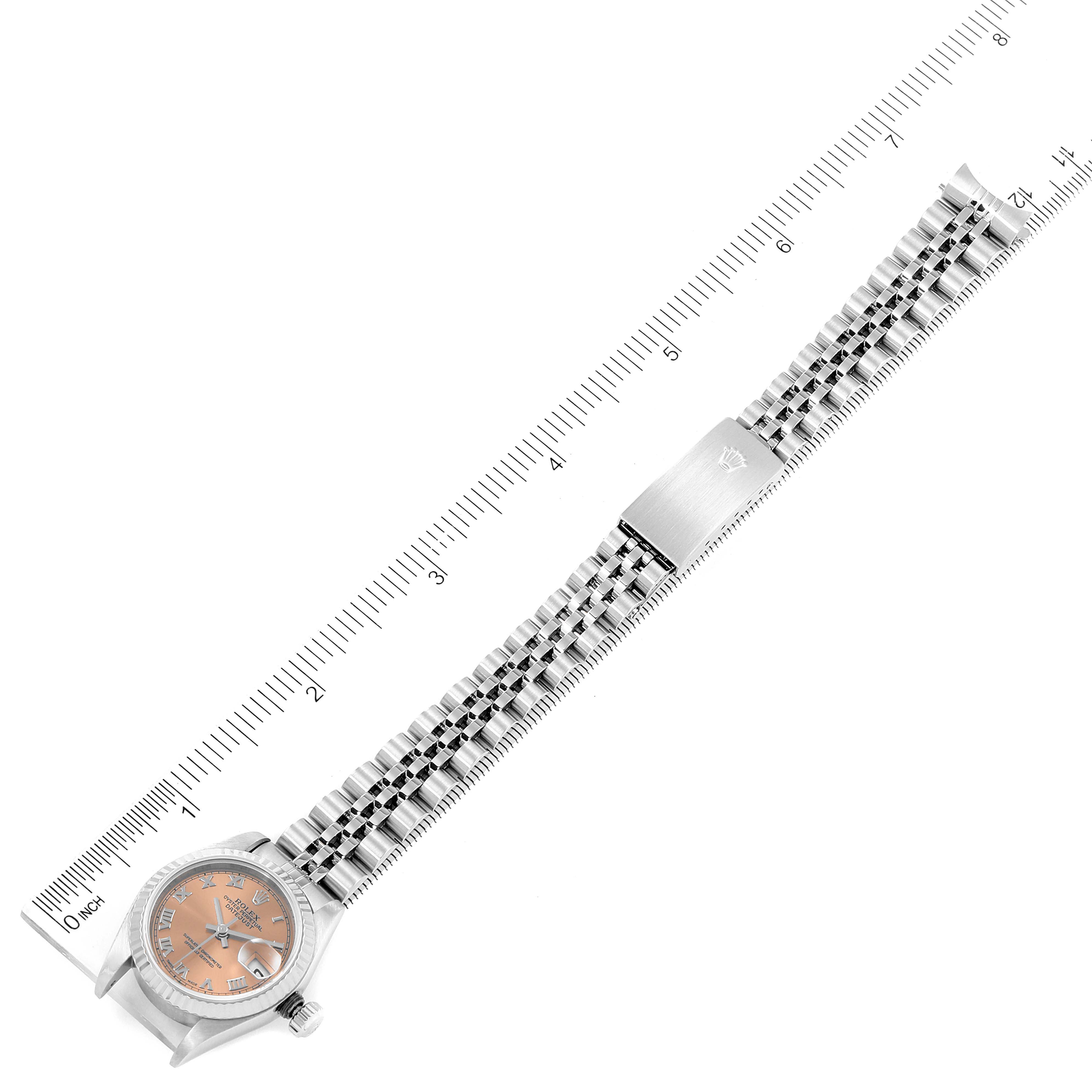 The image shows a top-down view of a Rolex Datejust 79174 Women's Stainless Steel Pink Dial 79174 Women's Stainless Steel Pink Dial watch with a metal bracelet and an orange dial, placed alongside a ruler.