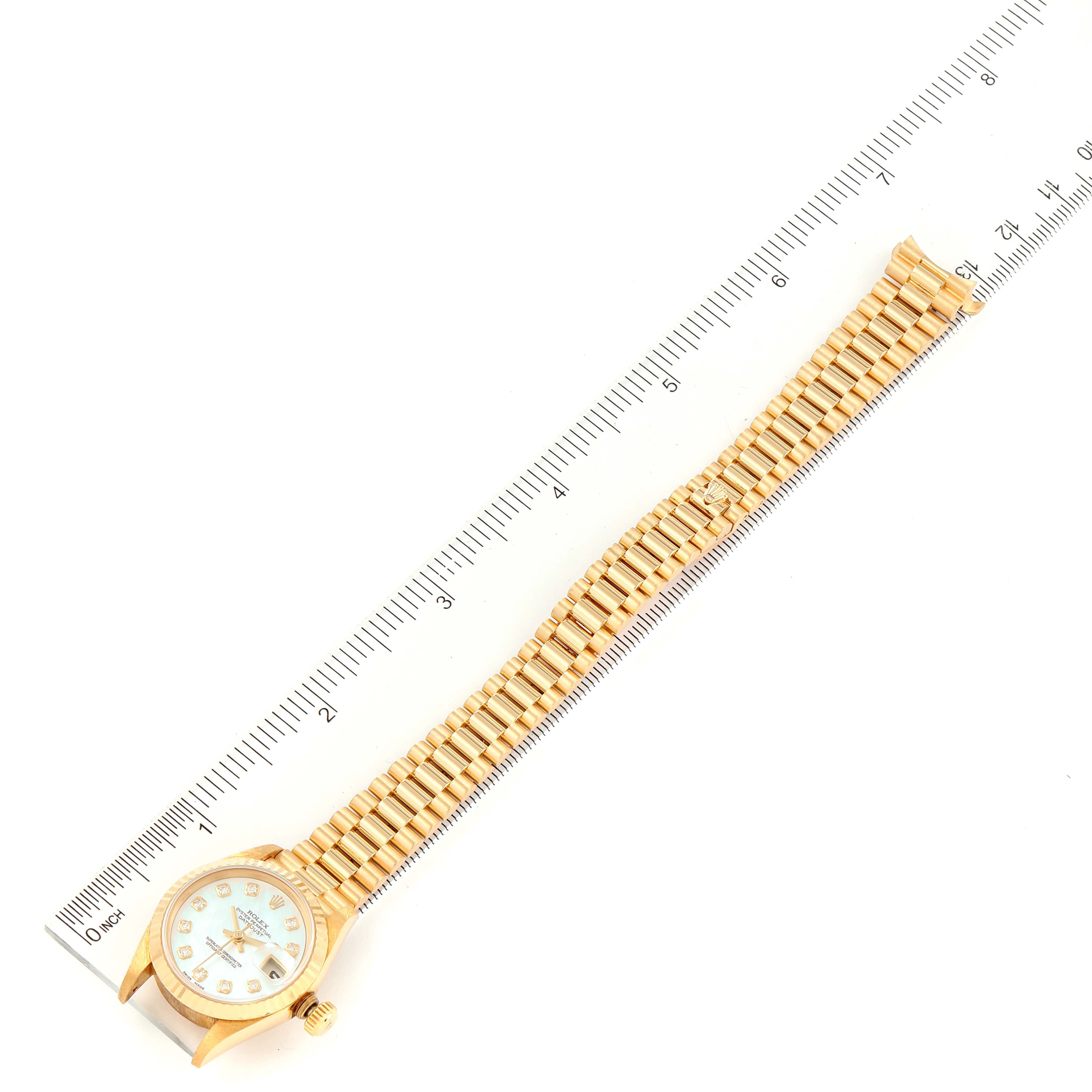 The image shows a Rolex President 69178 Women
s Yellow Gold Mother of Pearl Dial 69178 Women
s Yellow Gold Mother of Pearl Dial model watch from a top-down angle with its band fully extended next to a ruler.