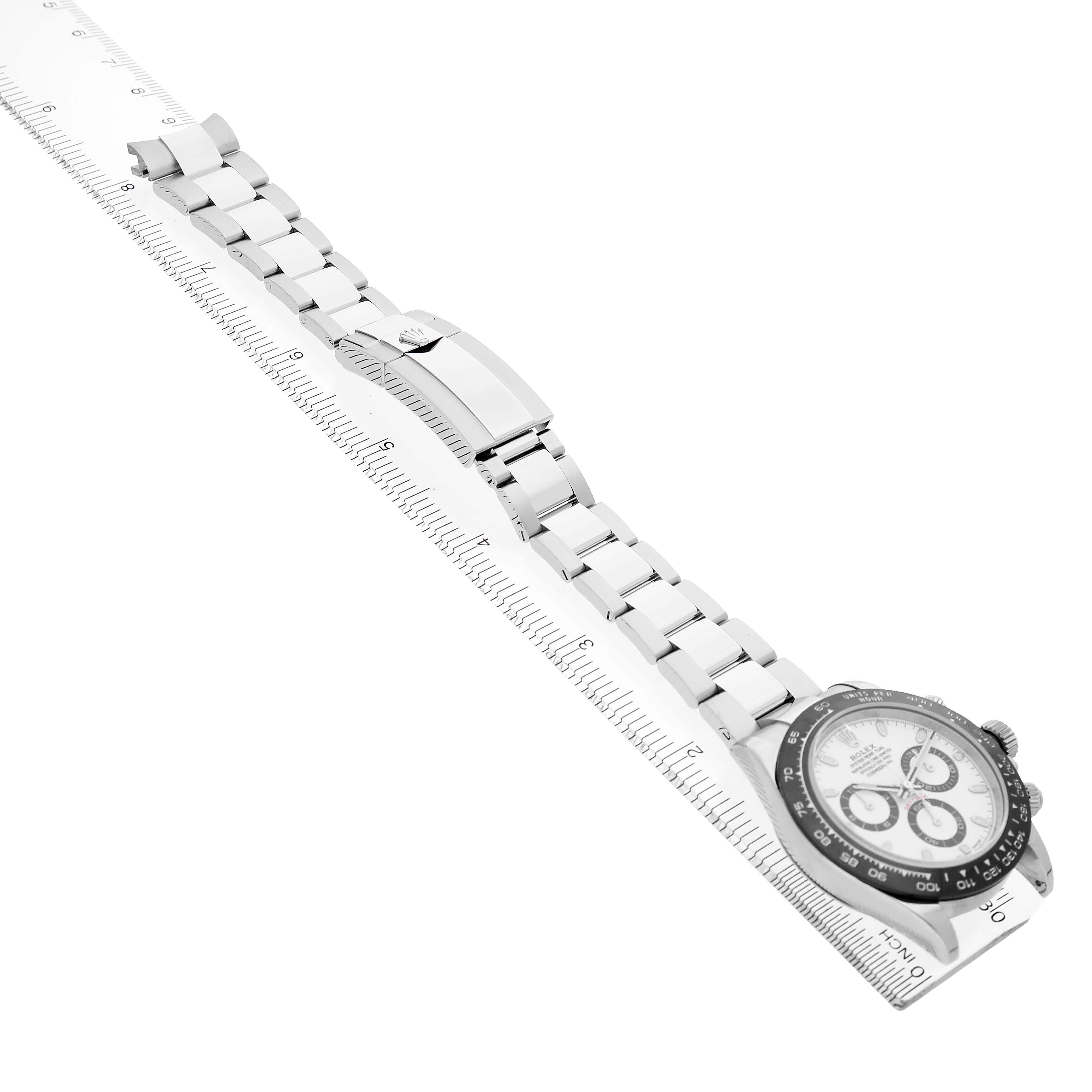 The Rolex Daytona M116500LN-0001 Men's Stainless Steel White Dial watch is shown in a side view, highlighting the bracelet, clasp, and watch face on a ruler.