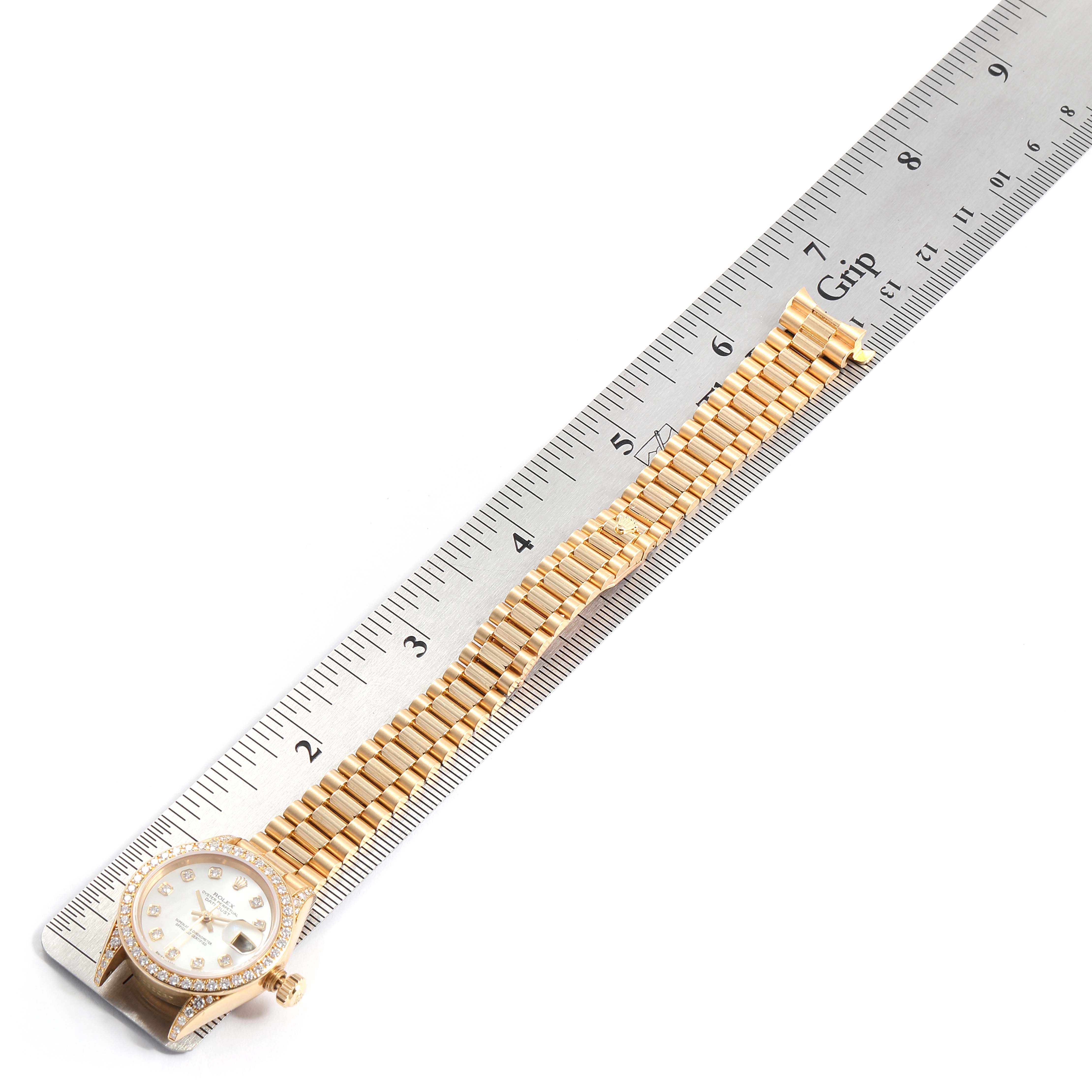 The image shows a Rolex President 69158 Women
s Yellow Gold Mother of Pearl Dial 69158 Women
s Yellow Gold Mother of Pearl Dial model watch laid next to a ruler, highlighting the bracelet and face of the watch.