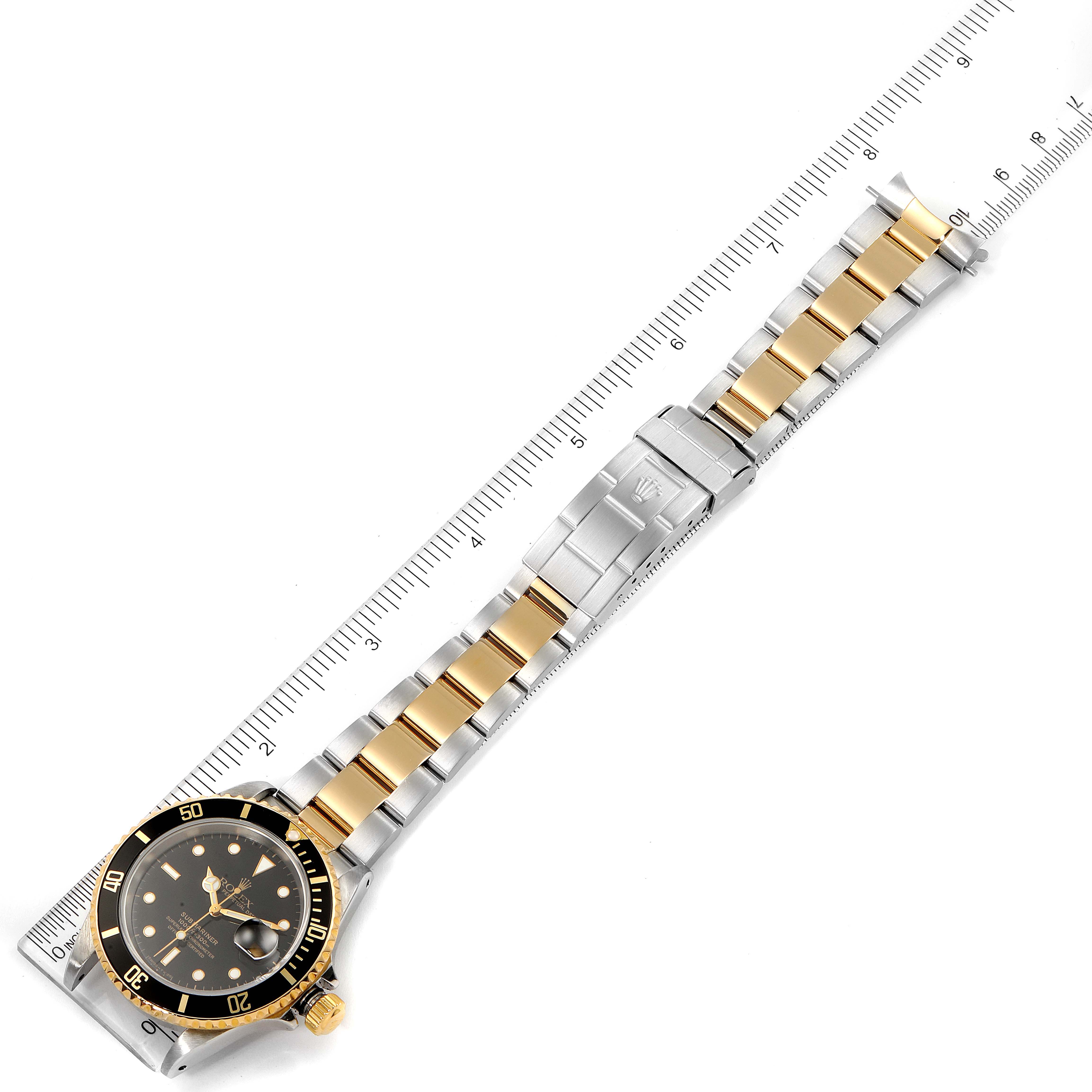The image shows a Rolex Submariner 16613 Men's Steel and Gold (two tone) Black Dial 16613 Men's Steel and Gold (two tone) Black Dial watch laid flat alongside a ruler, displaying its gold and silver bracelet and black bezel from an overhead view.