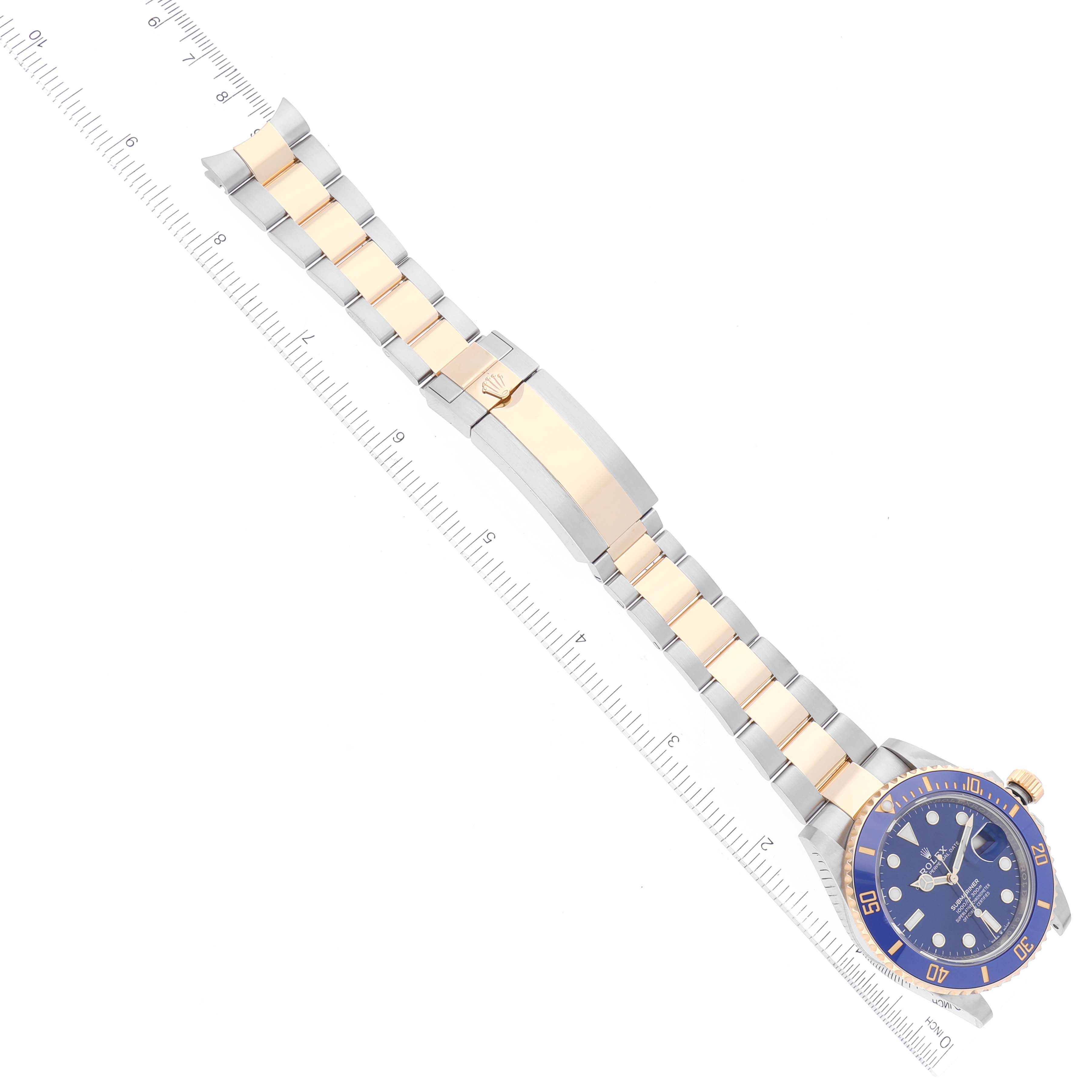 The image shows a Rolex Submariner 126613 Men's Steel and Gold (two tone) Blue Dial 126613 Men's Steel and Gold (two tone) Blue Dial watch laid flat with its blue bezel, dial, and two-tone metal bracelet visible alongside a ruler.