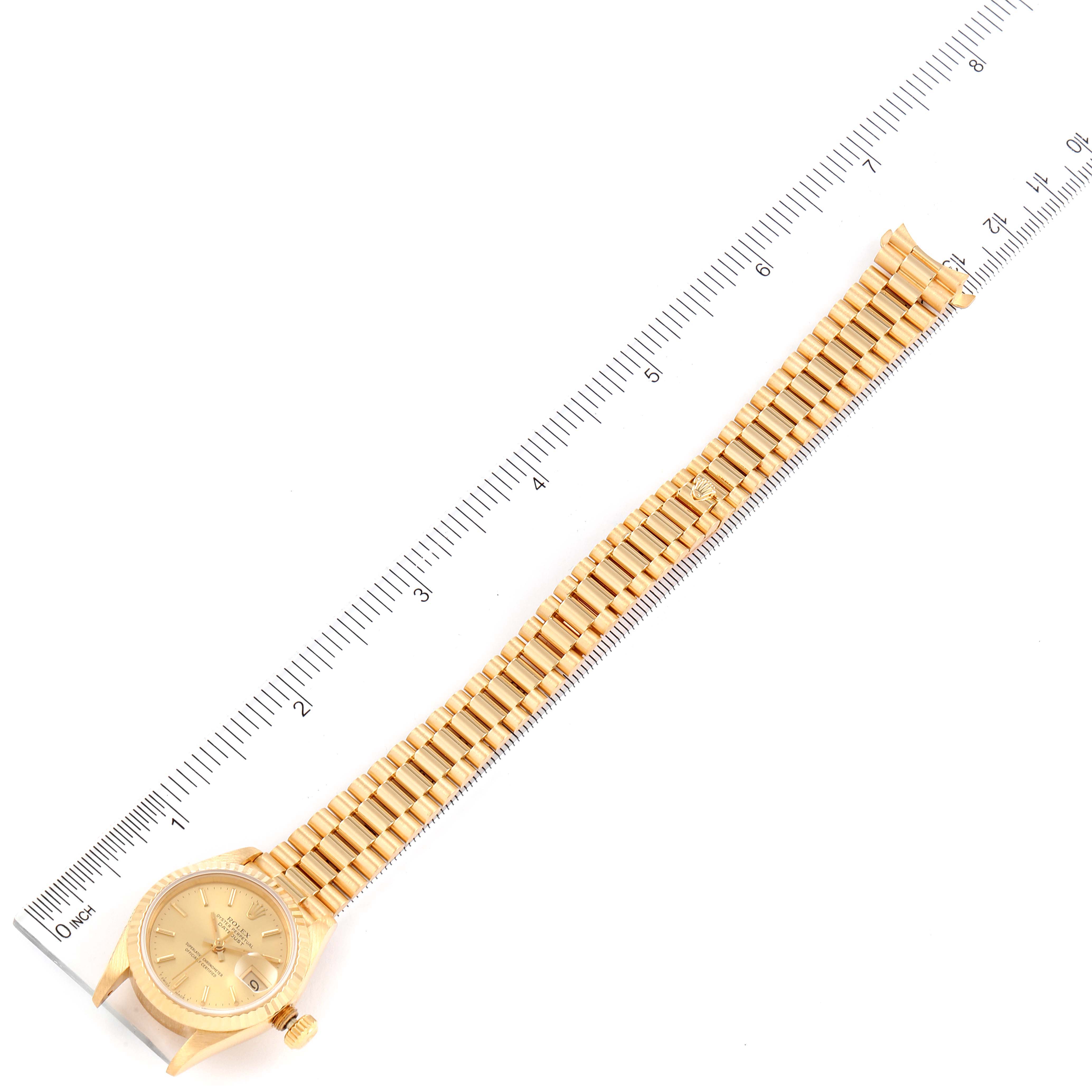 The image shows a Rolex President 69178 Women
s Yellow Gold Champagne Dial 69178 Women
s Yellow Gold Champagne Dial model watch laid flat next to a ruler, showcasing its entire band and face from a top-down angle.