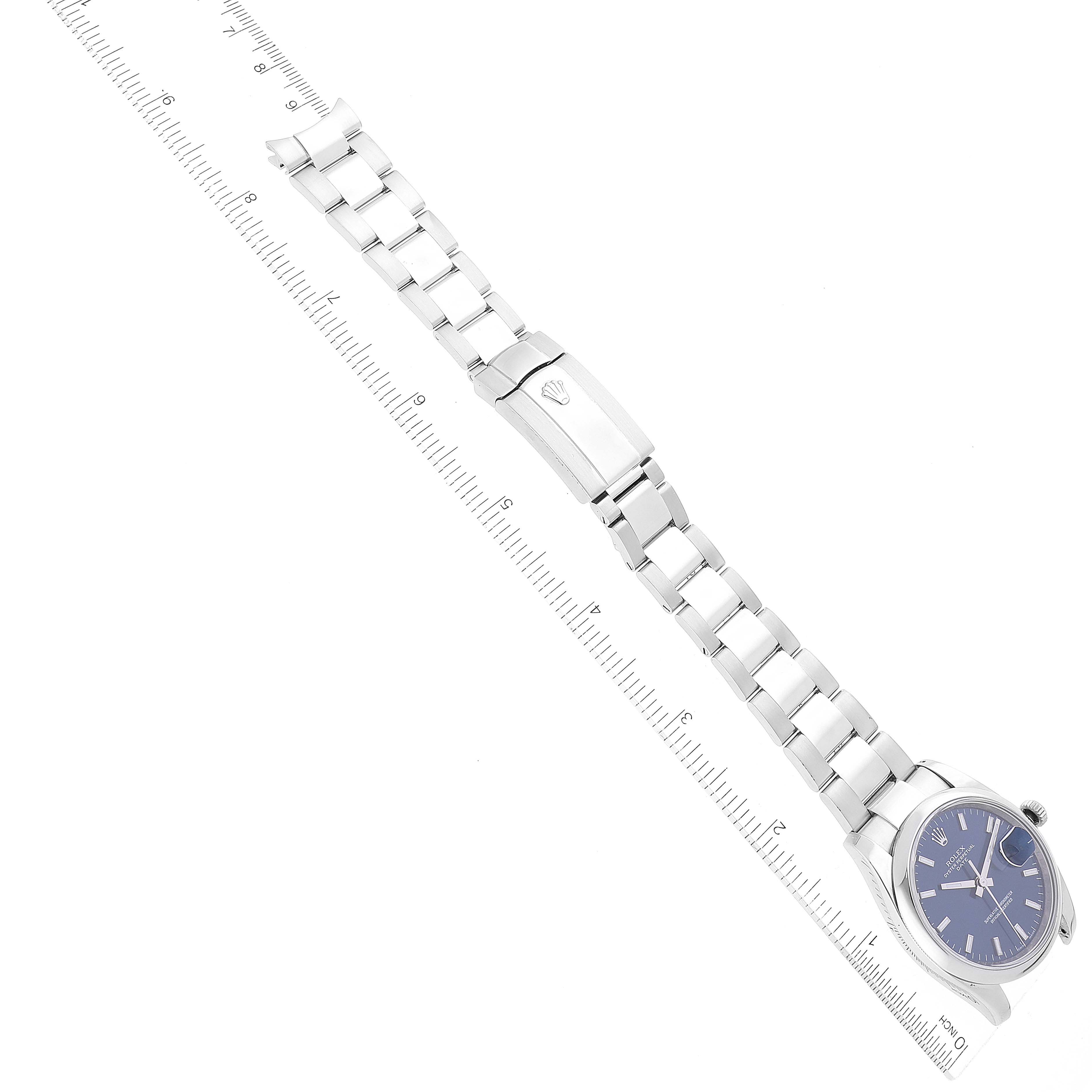 The image shows a Rolex Date 115200 Men's Stainless Steel Blue Dial 115200 Men's Stainless Steel Blue Dial model watch laid flat against a ruler, highlighting the bracelet and face.