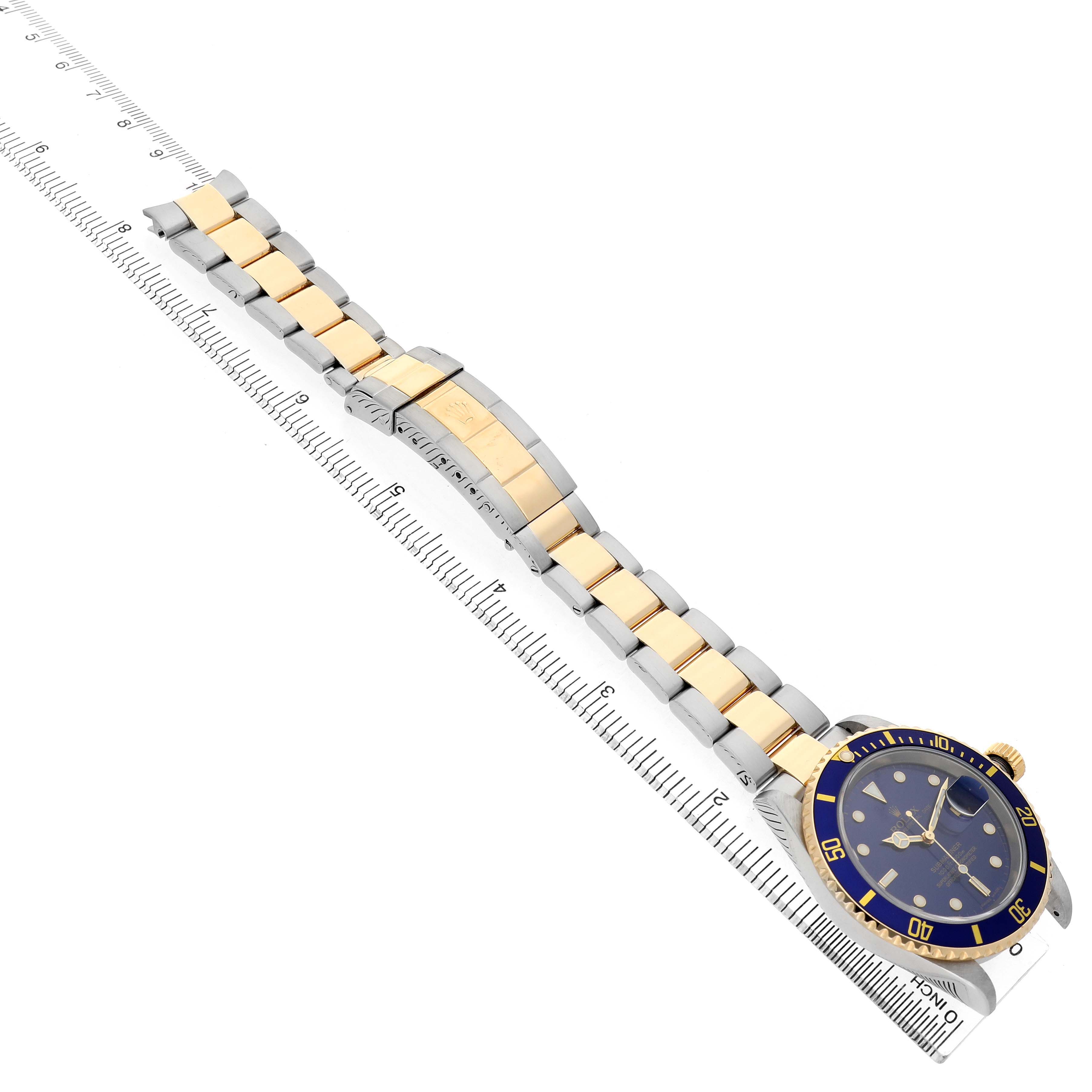 The Rolex Submariner 16613 T Men's Steel and Gold (two tone) Blue Dial is shown at an angle displaying the full watch and bracelet, with a ruler alongside for scale.
