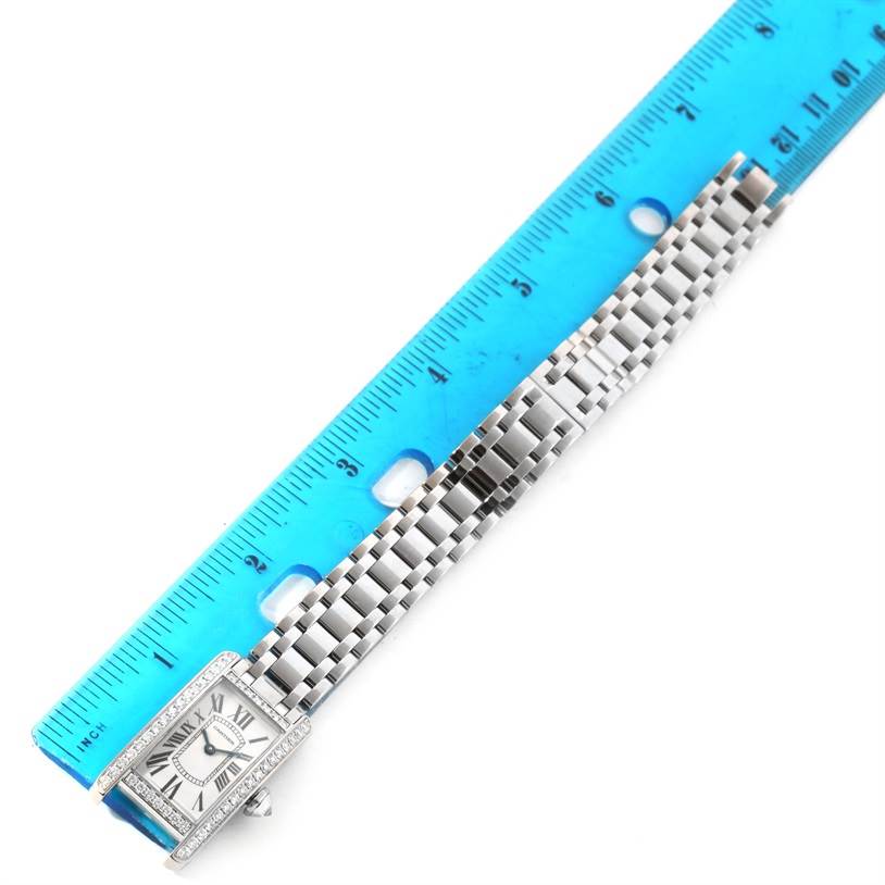 The image shows the Cartier Tank Americaine WB7073L1 Women's White Gold Silver Dial WB7073L1 Women's White Gold Silver Dial watch laid flat with its rectangular face and stainless steel strap next to a ruler.