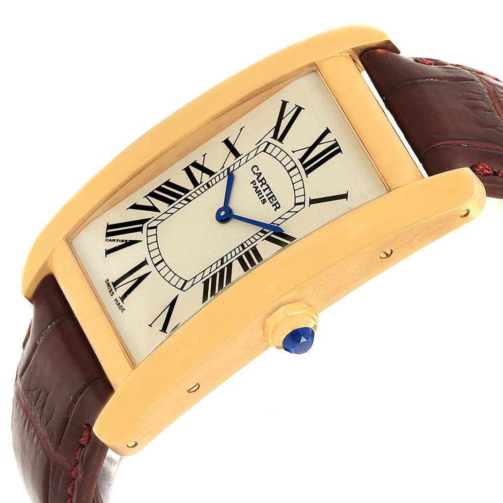 The image shows a side angle of the Cartier Tank Americaine 1735 Men
s Yellow Gold Silver Dial 1735 Men
s Yellow Gold Silver Dial watch, highlighting its gold case, Roman numerals, blue hands, and leather strap.