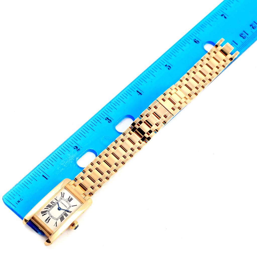 The image shows a Tank Américaine model by Cartier laid flat next to a blue ruler, highlighting the watch face and metal bracelet.