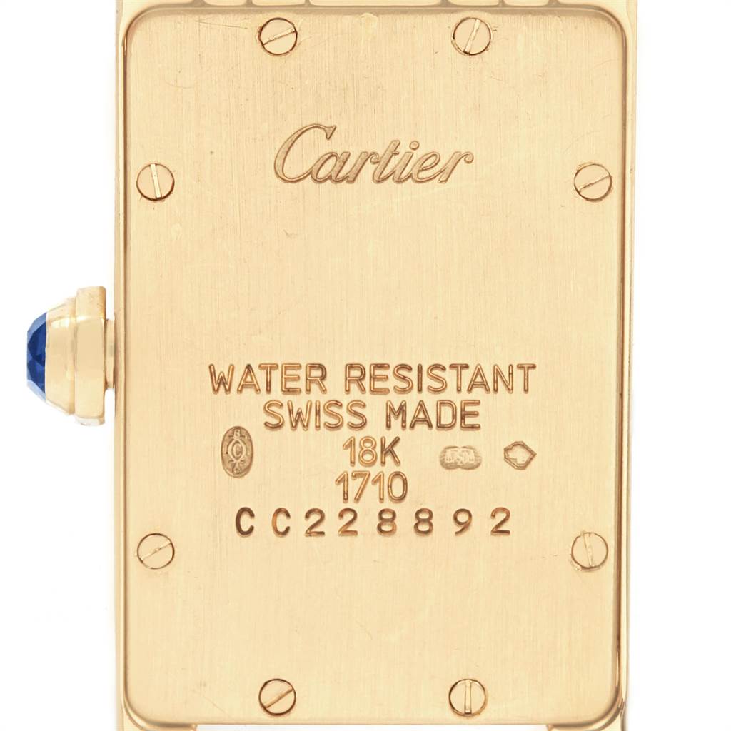 The image shows the back view of a Tank Americaine watch by Cartier, displaying inscriptions and the crown with a gem.