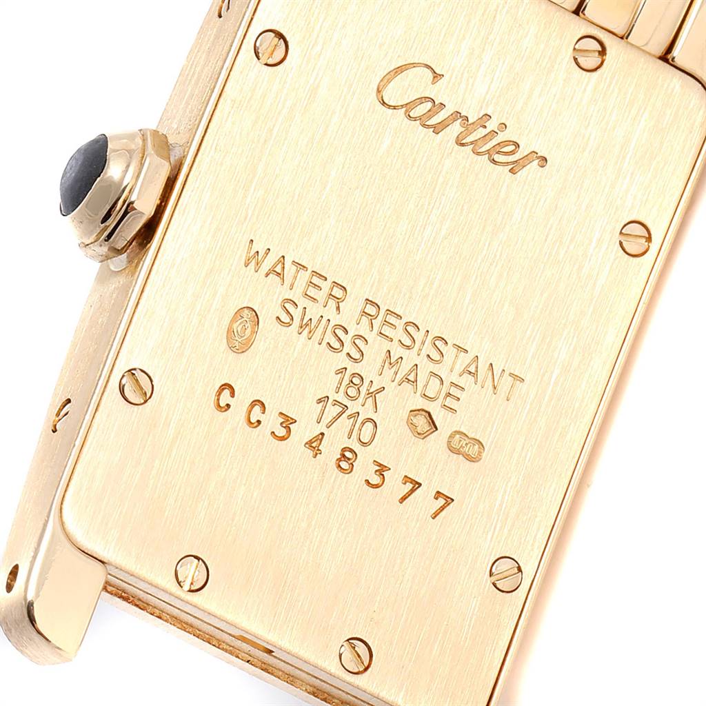 The image shows the back case of a Cartier Tank Americaine W26015K2 Women
s Yellow Gold Silver Dial W26015K2 Women
s Yellow Gold Silver Dial watch, highlighting engraved details and a crown.