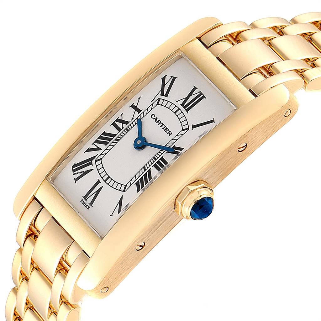 The image shows a Cartier Tank Americaine W26015K2 Women's Yellow Gold Silver Dial W26015K2 Women's Yellow Gold Silver Dial watch at an angle highlighting its gold case, blue hands, Roman numerals, and sapphire crown.