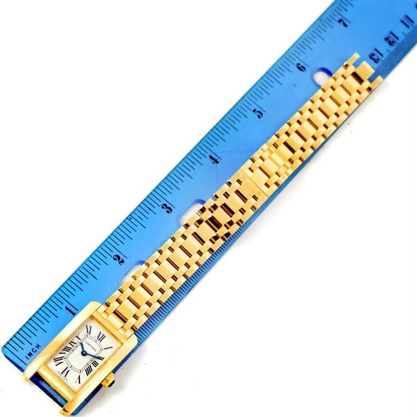 This image shows a Cartier Tank Americaine W26015K2 Women
s Yellow Gold Silver Dial W26015K2 Women
s Yellow Gold Silver Dial watch placed next to a blue ruler, displaying the face and gold bracelet.